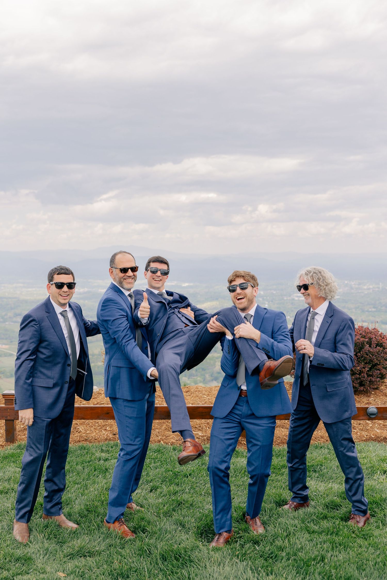 Groomsmen in navy suits and sunglasses playfully lifting groom on grass with mountain backdrop.