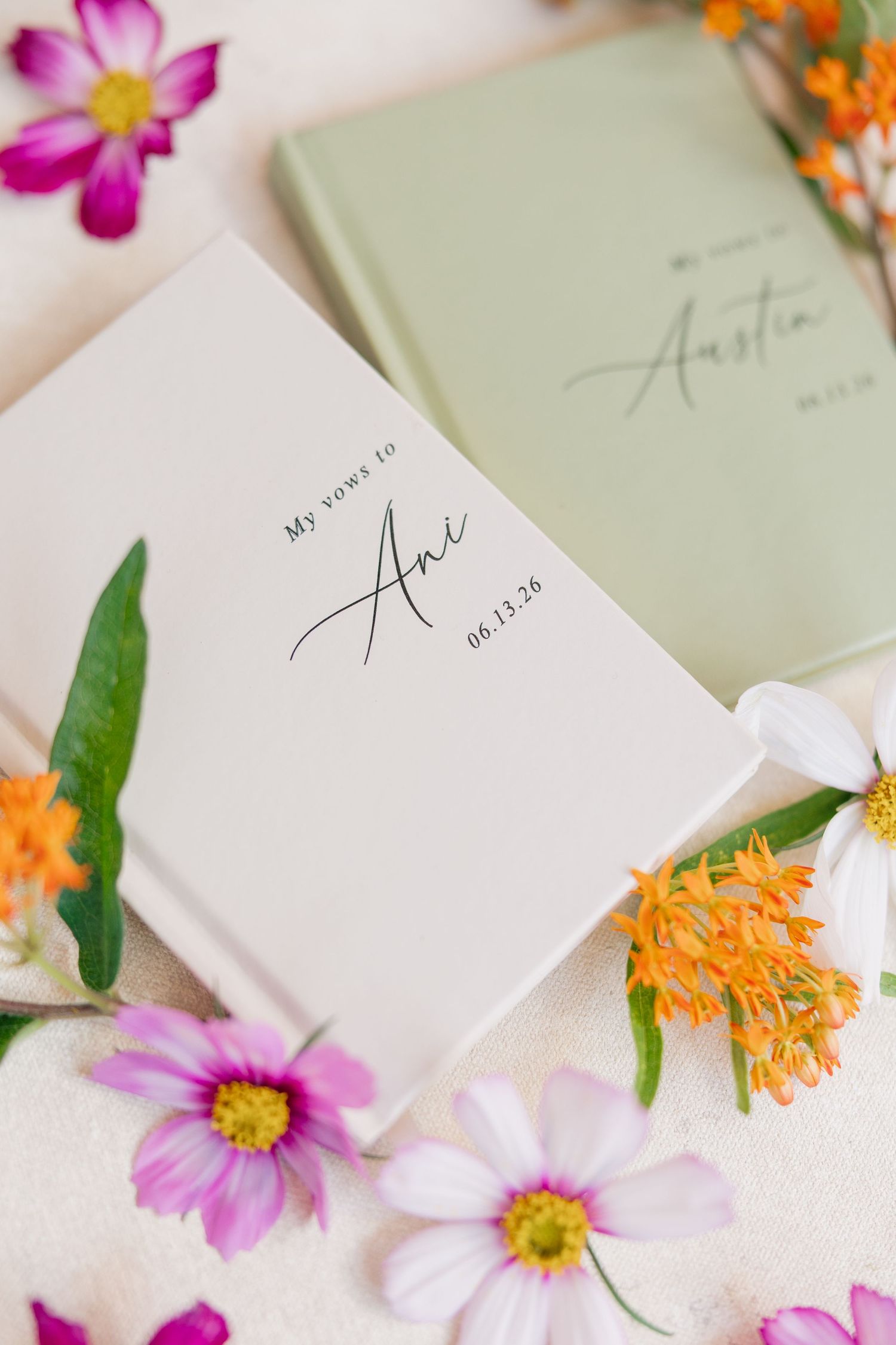 Two personalized white and green wedding vow books surrounded by colorful wildflowers including pink cosmos and orange blooms.