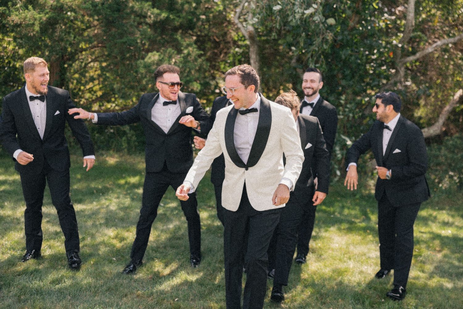 Groom in white tuxedo jacket laughing with groomsmen in black tuxedos outdoors on green grass.