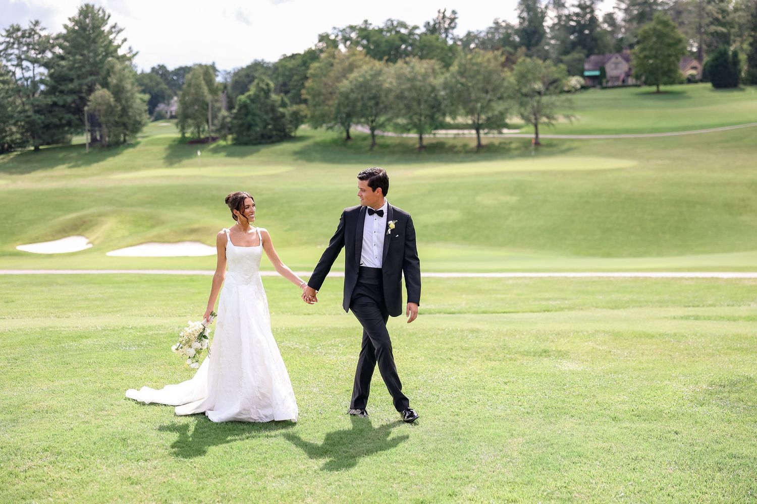 Bride and groom walking hand in hand across lush green golf course on their wedding day.