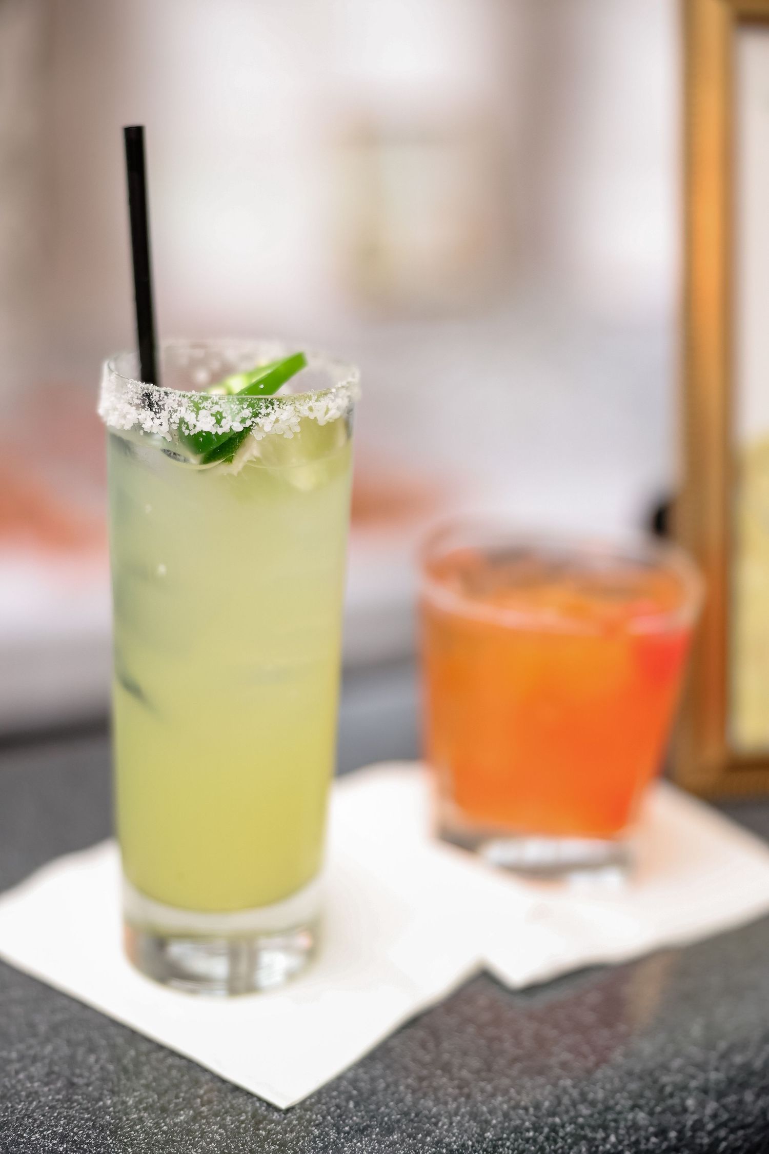 Two cocktails on a bar, a yellow margarita with salted rim and black straw, beside an orange drink on napkins.