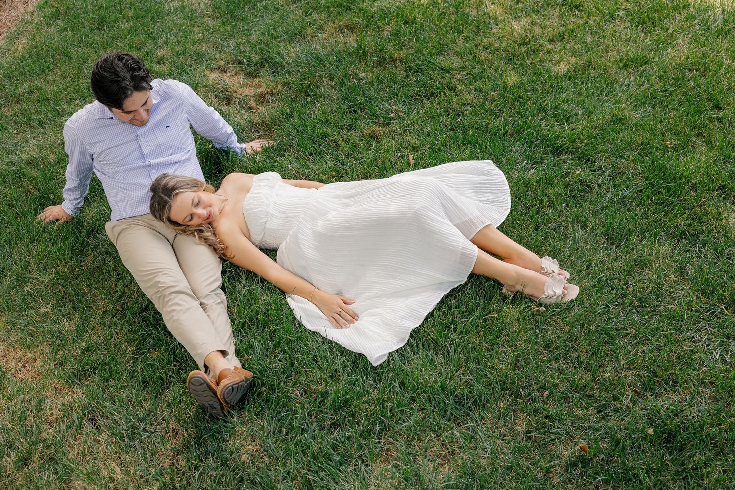 Bride in white dress and groom in khakis laying on green grass together during outdoor engagement photo session.