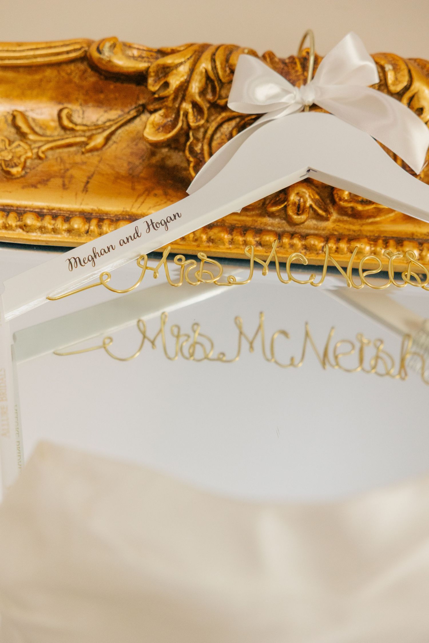 Custom white wedding hanger with gold Mrs. McNetsh wire lettering and white bow leaning against ornate gold mirror.