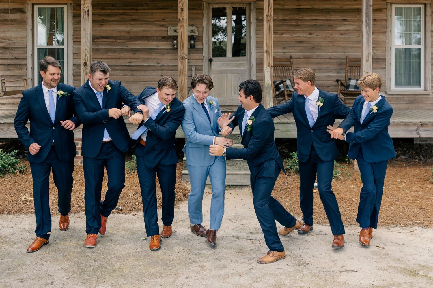 Groom in light blue suit laughs with six groomsmen in navy suits outside a rustic wooden building.