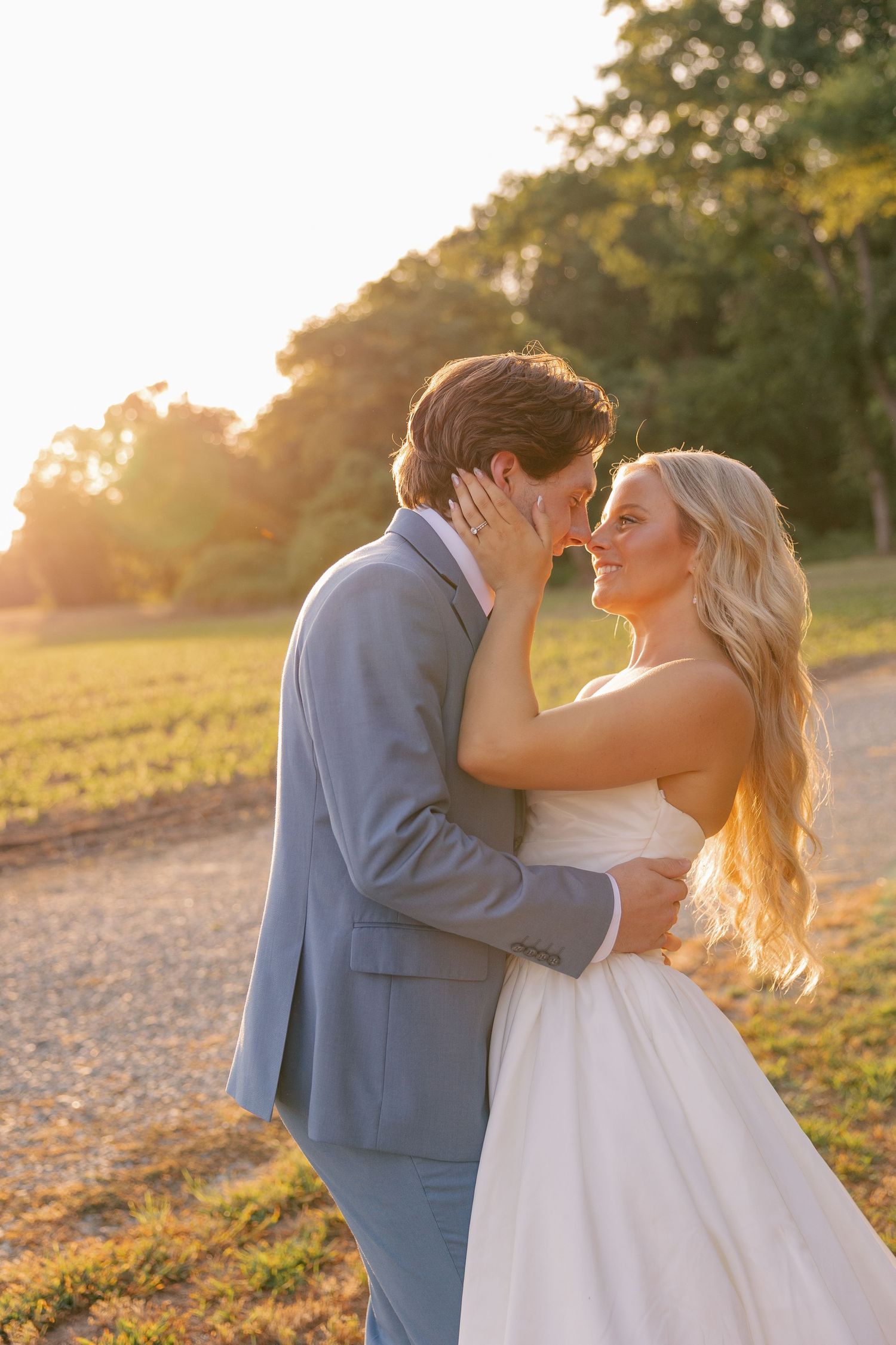 Golden hour wedding photo of bride and groom sharing a romantic kiss outdoors in a sunlit field.