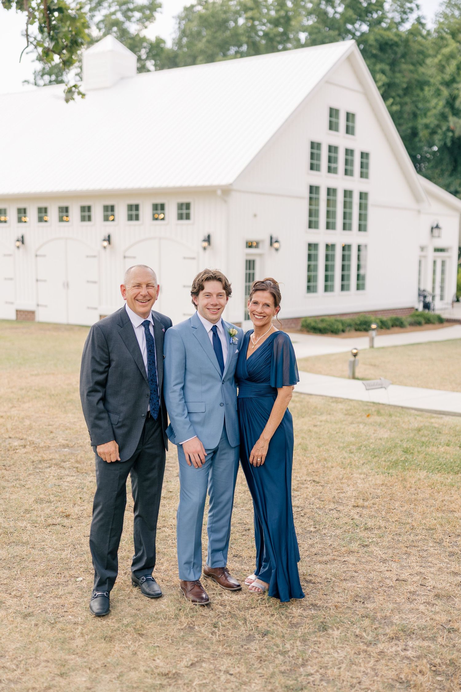 Groom in light blue suit poses with parents outside a white barn wedding venue on wedding day.