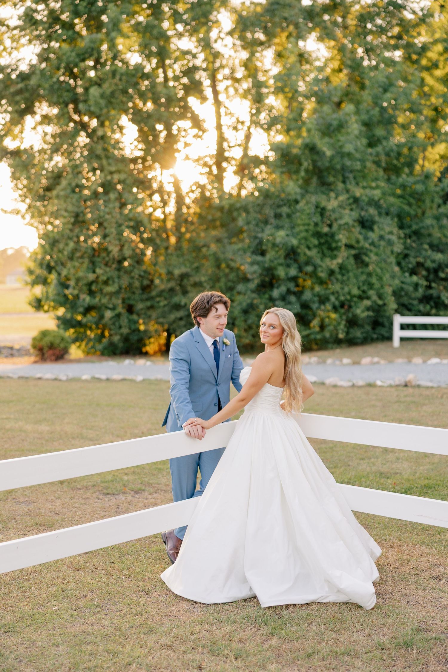 Bride in strapless white gown and groom in blue suit hold hands by white fence at golden hour sunset.