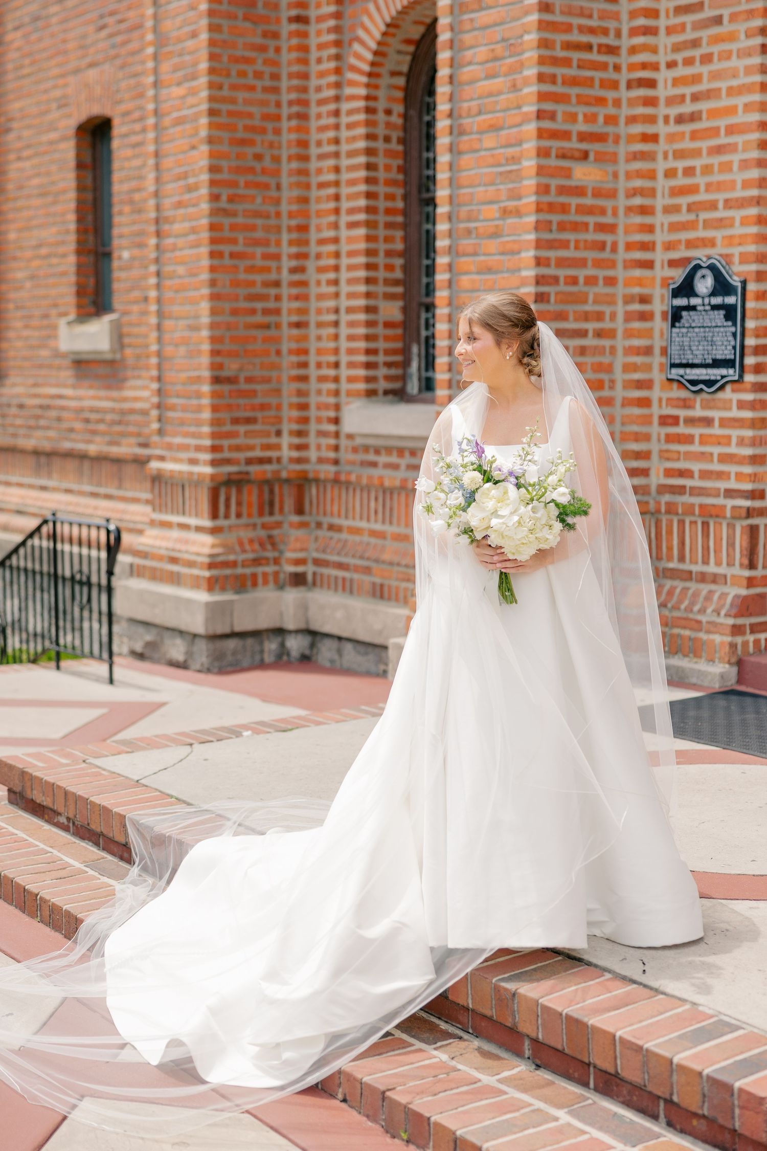 Bride in elegant white ball gown with cathedral veil holds bouquet of white flowers outside brick church.