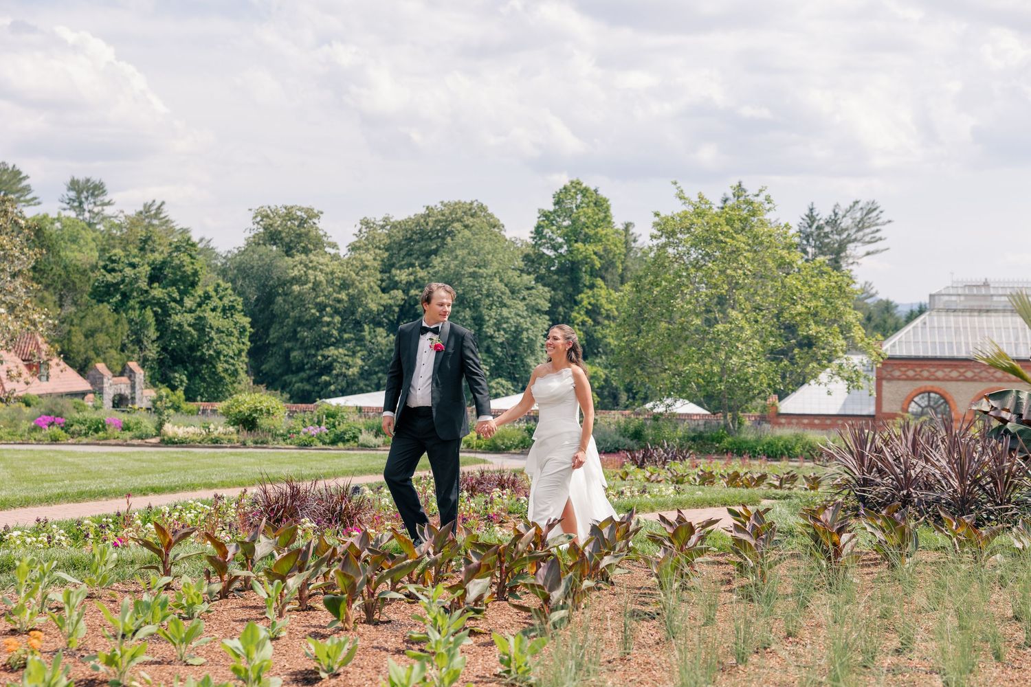 Bride and groom walk hand-in-hand through lush garden grounds on their summer wedding day.