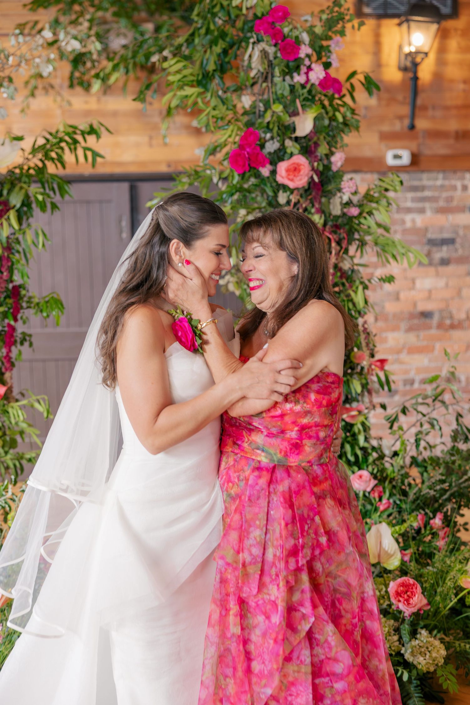Bride in white gown embracing smiling woman in pink floral dress before floral wedding arch backdrop.