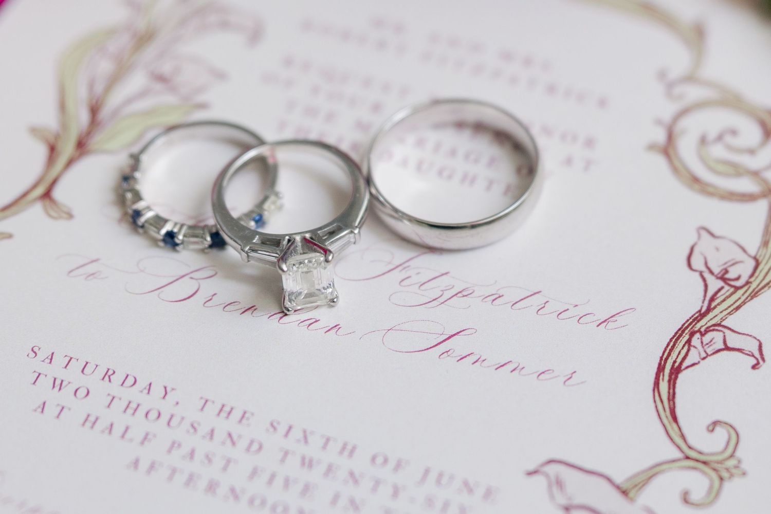 Three elegant wedding rings rest on a romantic wedding invitation with floral details and cursive script.
