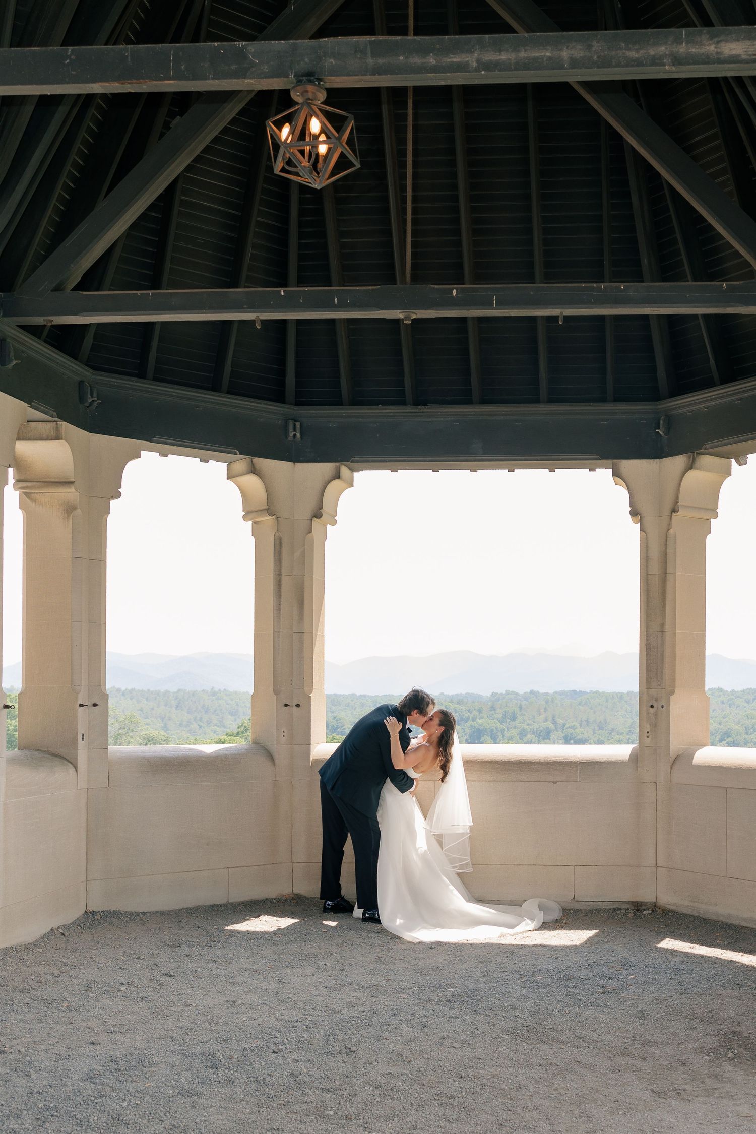 Bride and groom kiss inside an elegant open-air gazebo with stunning mountain landscape views behind them.