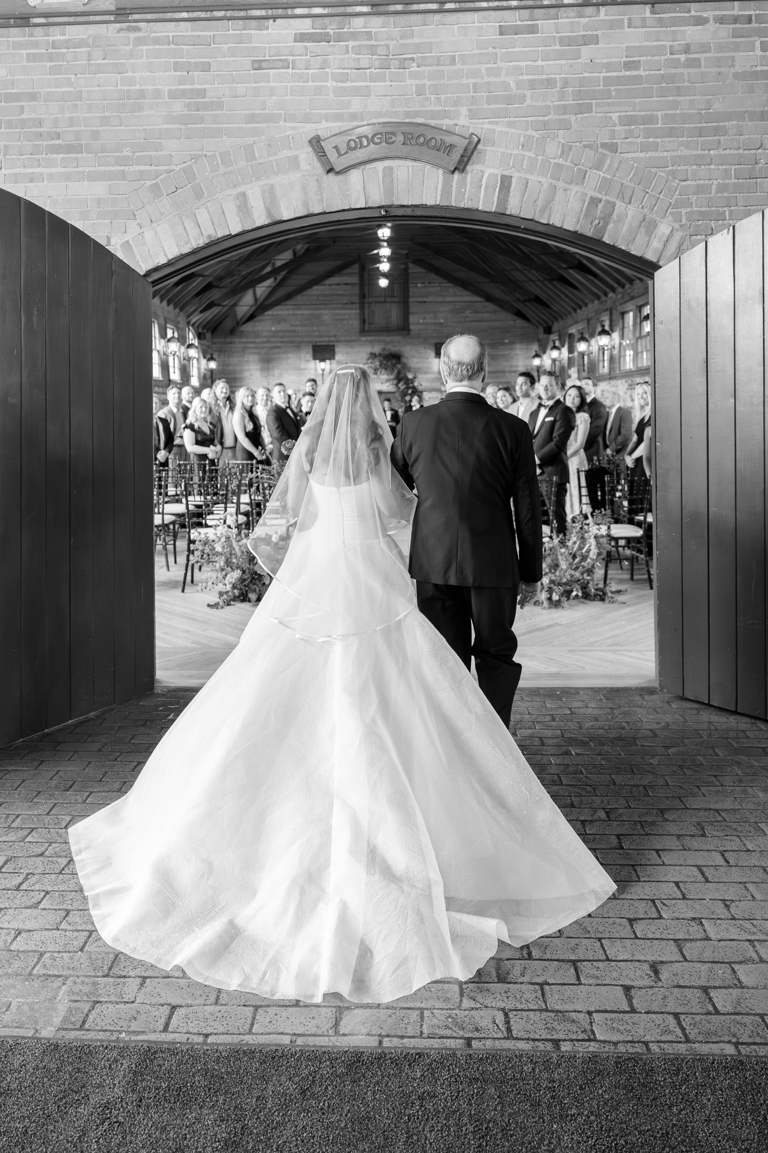 Black and white photo of bride in flowing gown walking down aisle with escort through brick archway.
