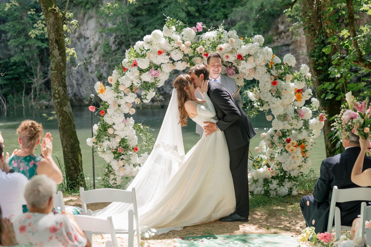 Bride and groom share first kiss under a stunning floral arch by a serene waterway during outdoor ceremony.