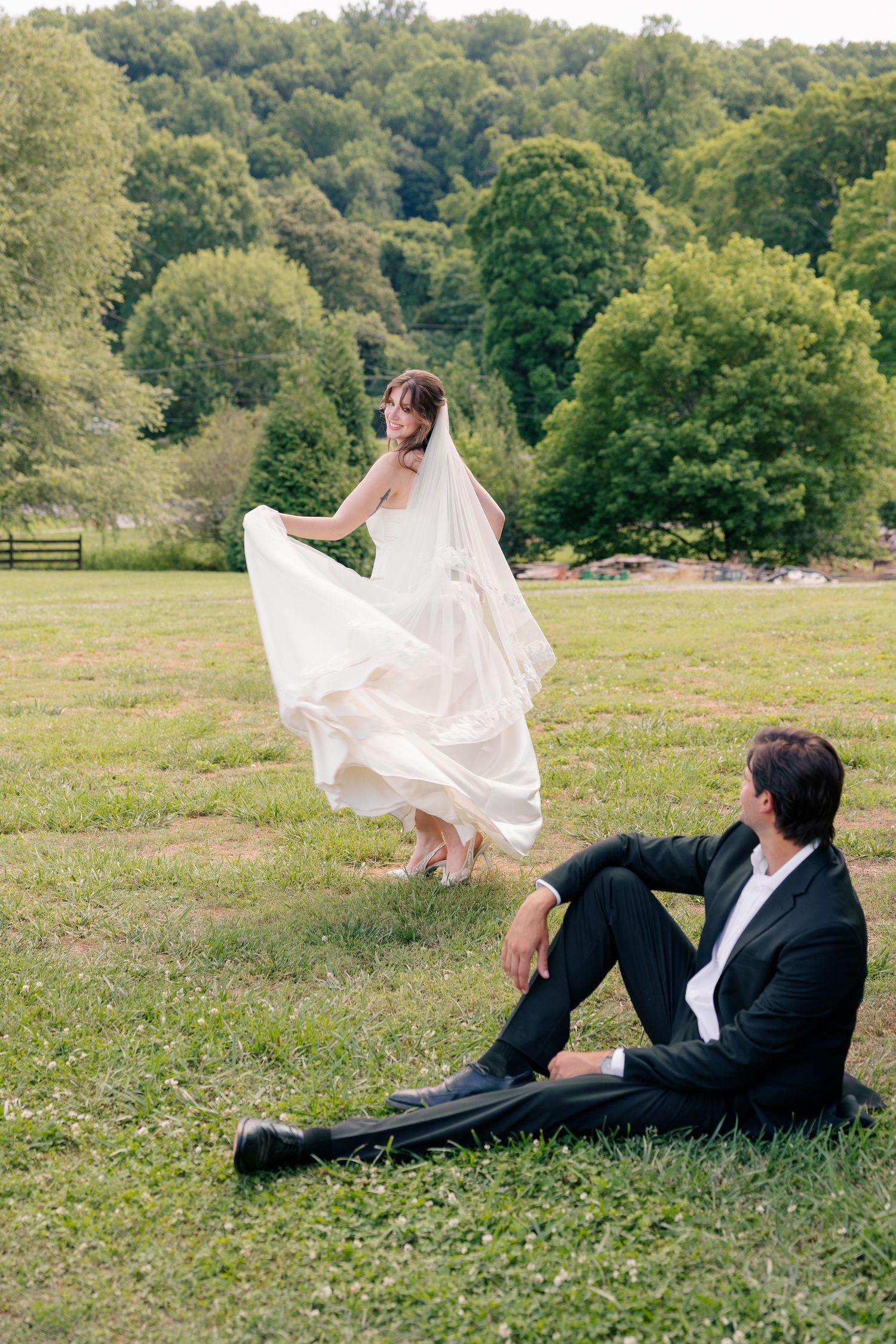 Bride twirling her gown in a green meadow while groom sits relaxed on the grass.