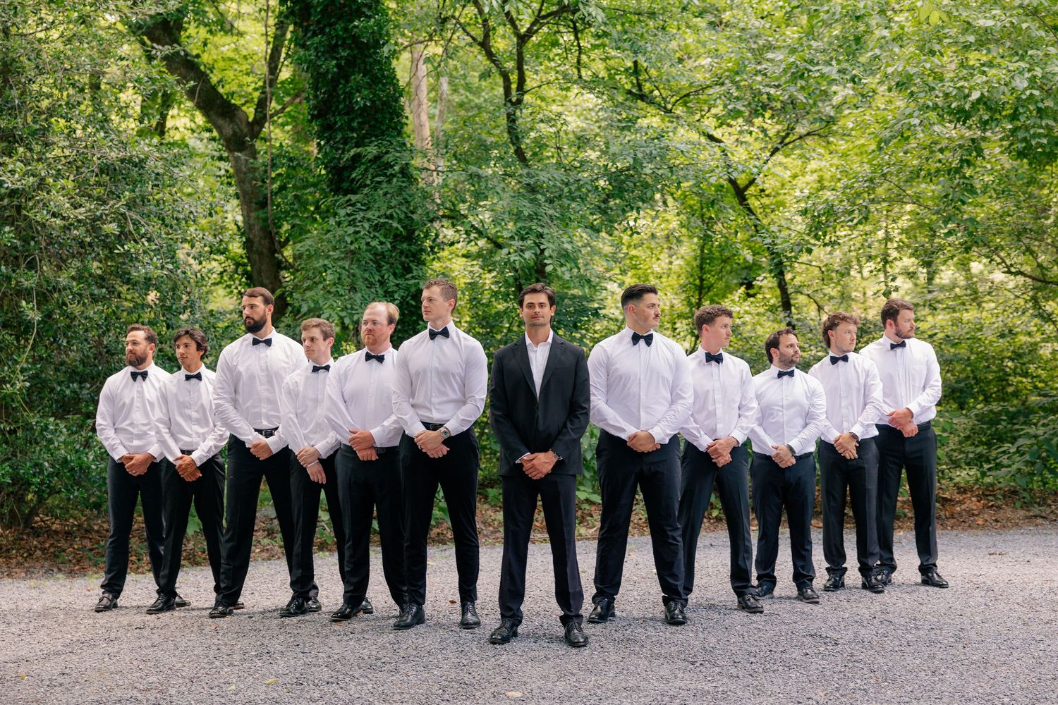 Groom stands with groomsmen in black tuxedos and white shirts on a tree-lined path.