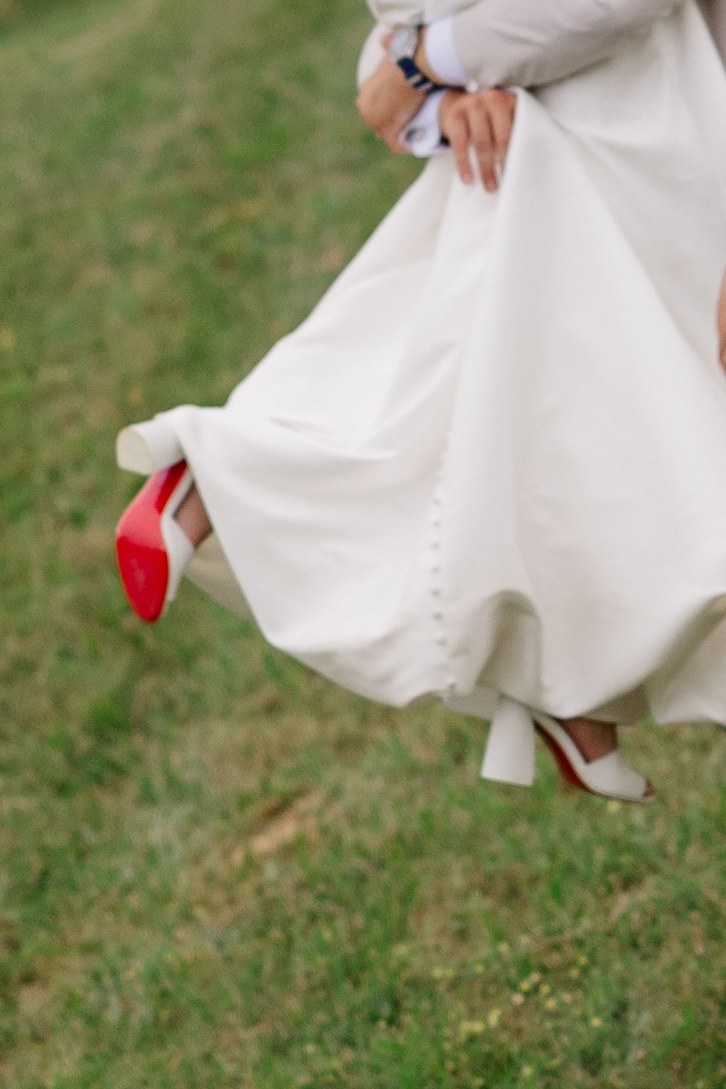 Close-up of a white wedding dress hem revealing a red-soled shoe on grass.