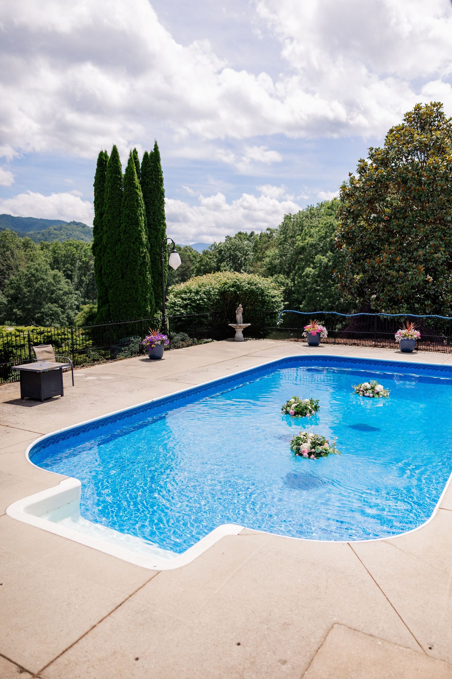 A luxurious swimming pool with floating floral arrangements surrounded by tall cypress trees and mountain views.