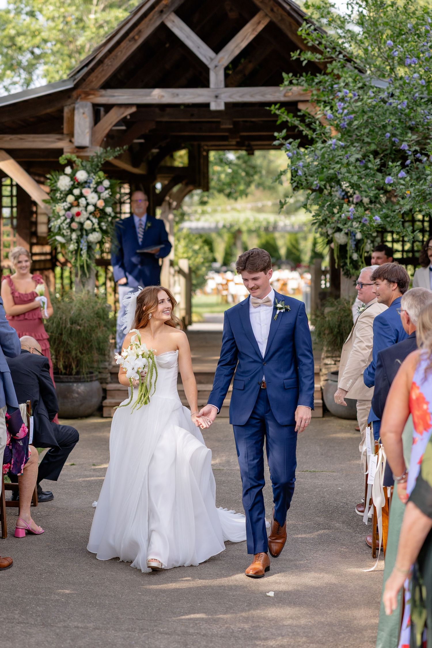 Wedding couple walks under a rustic wooden pavilion while guests throw flower petals in celebration.