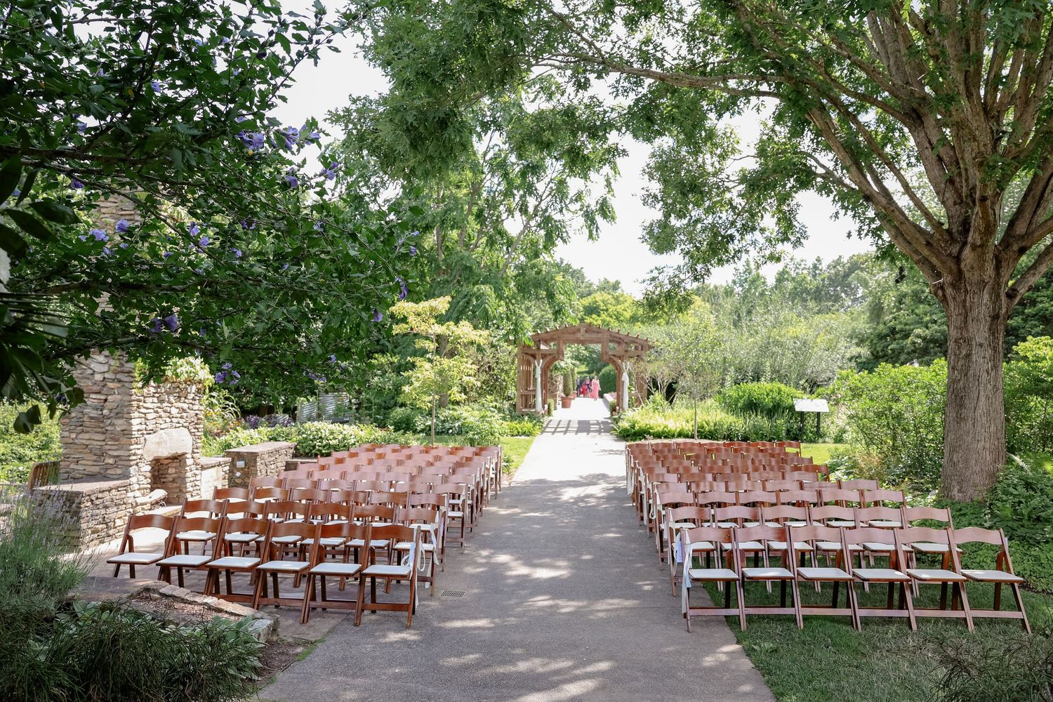 Wooden chairs line a garden pathway leading to a wedding ceremony arch.