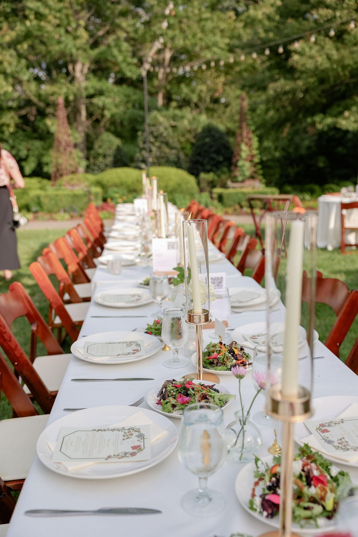 An elegant outdoor wedding table setting with white linens, candles and red chairs in a garden.