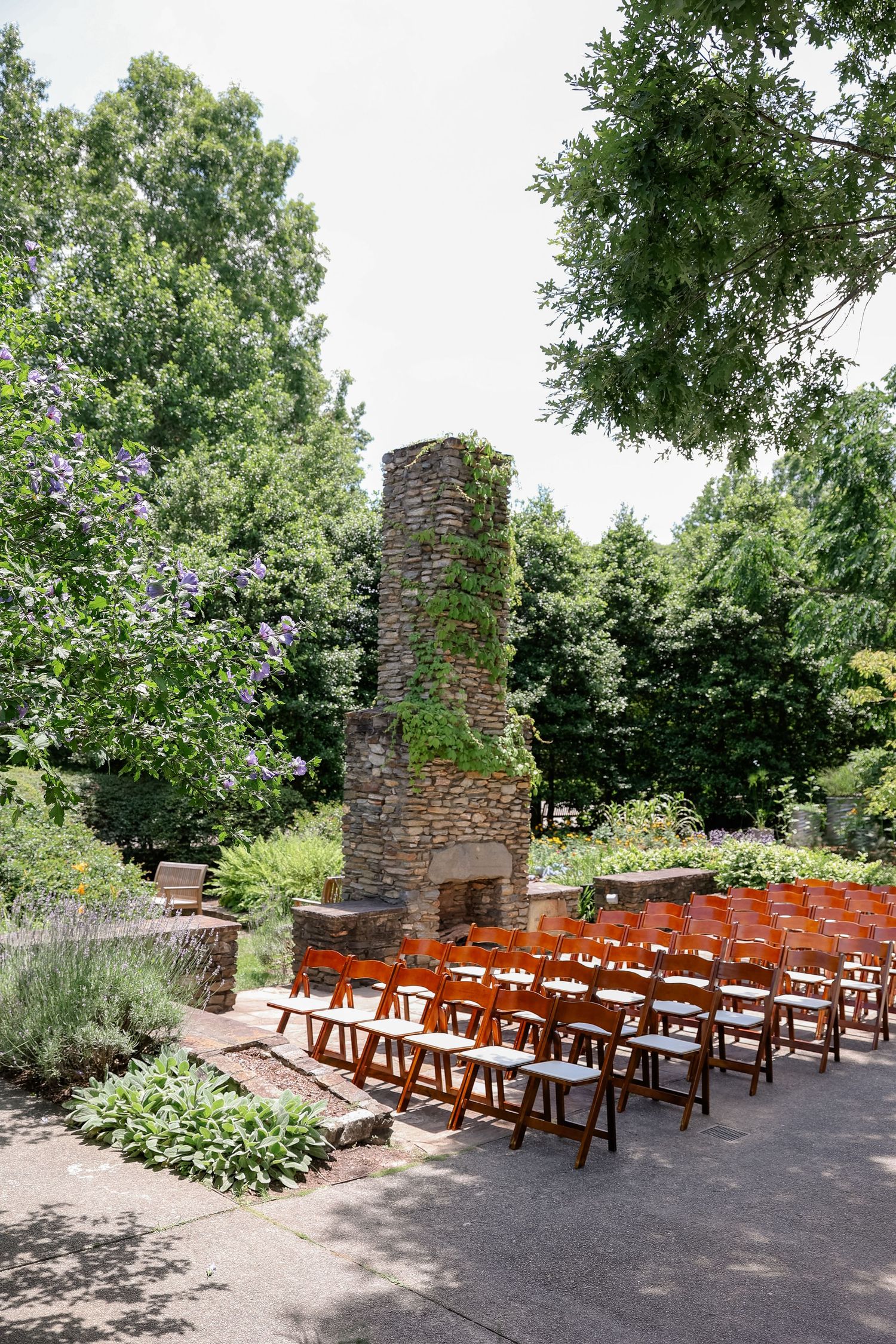 Outdoor wedding ceremony setup with wooden chairs and stone fireplace in a garden setting.