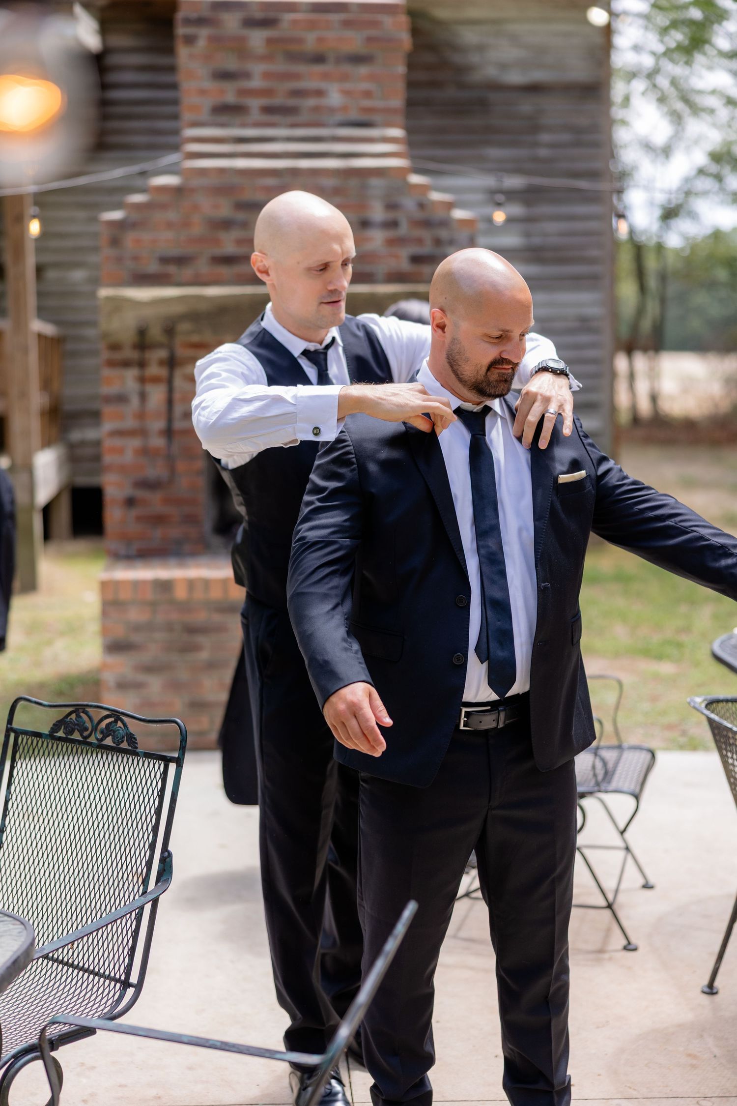 Two people in formal black suits playfully dance together on an outdoor patio with brick walls.