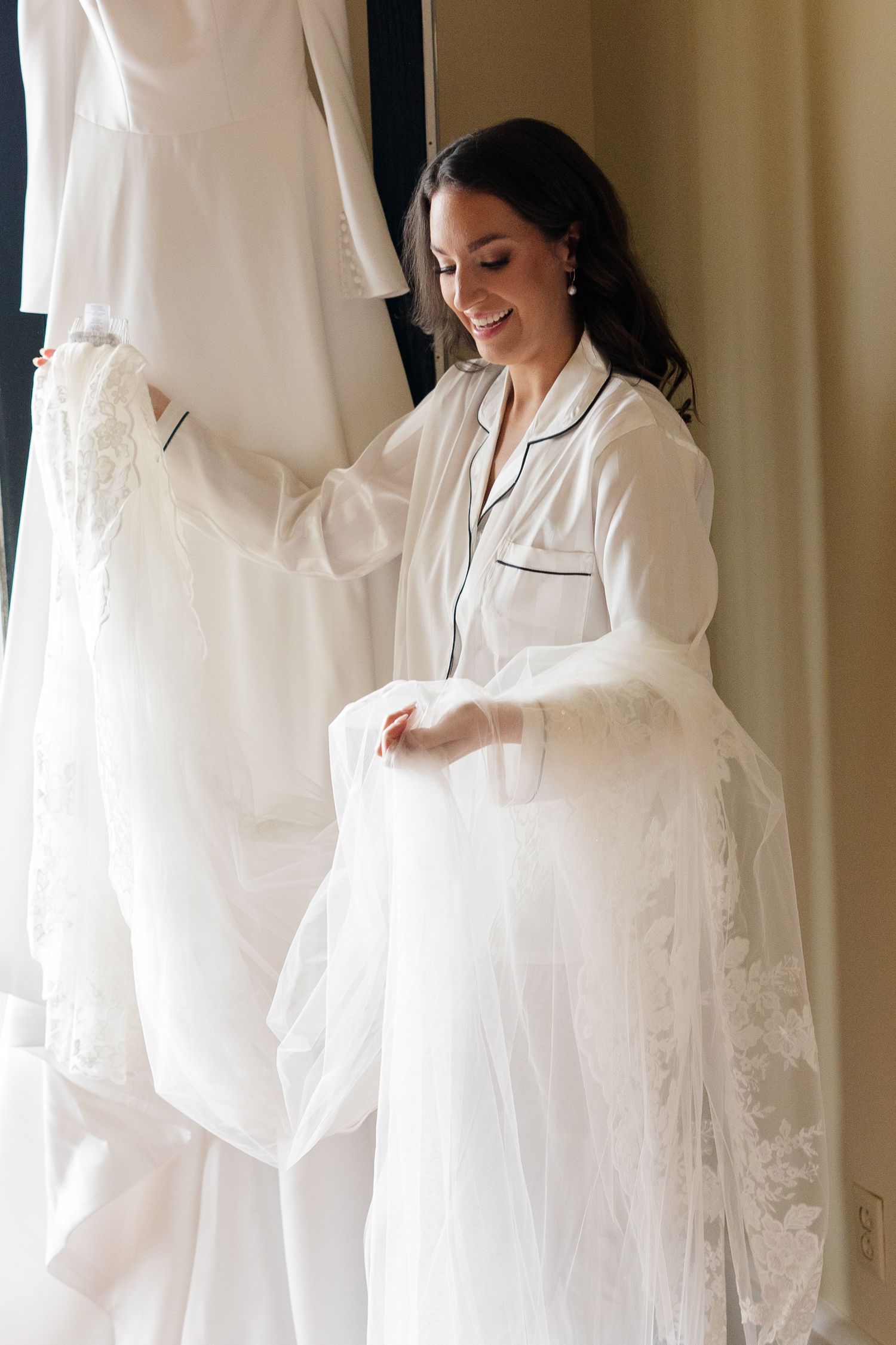 White wedding dress hanging in sunlit room while someone in white silk pajamas looks through clothing.