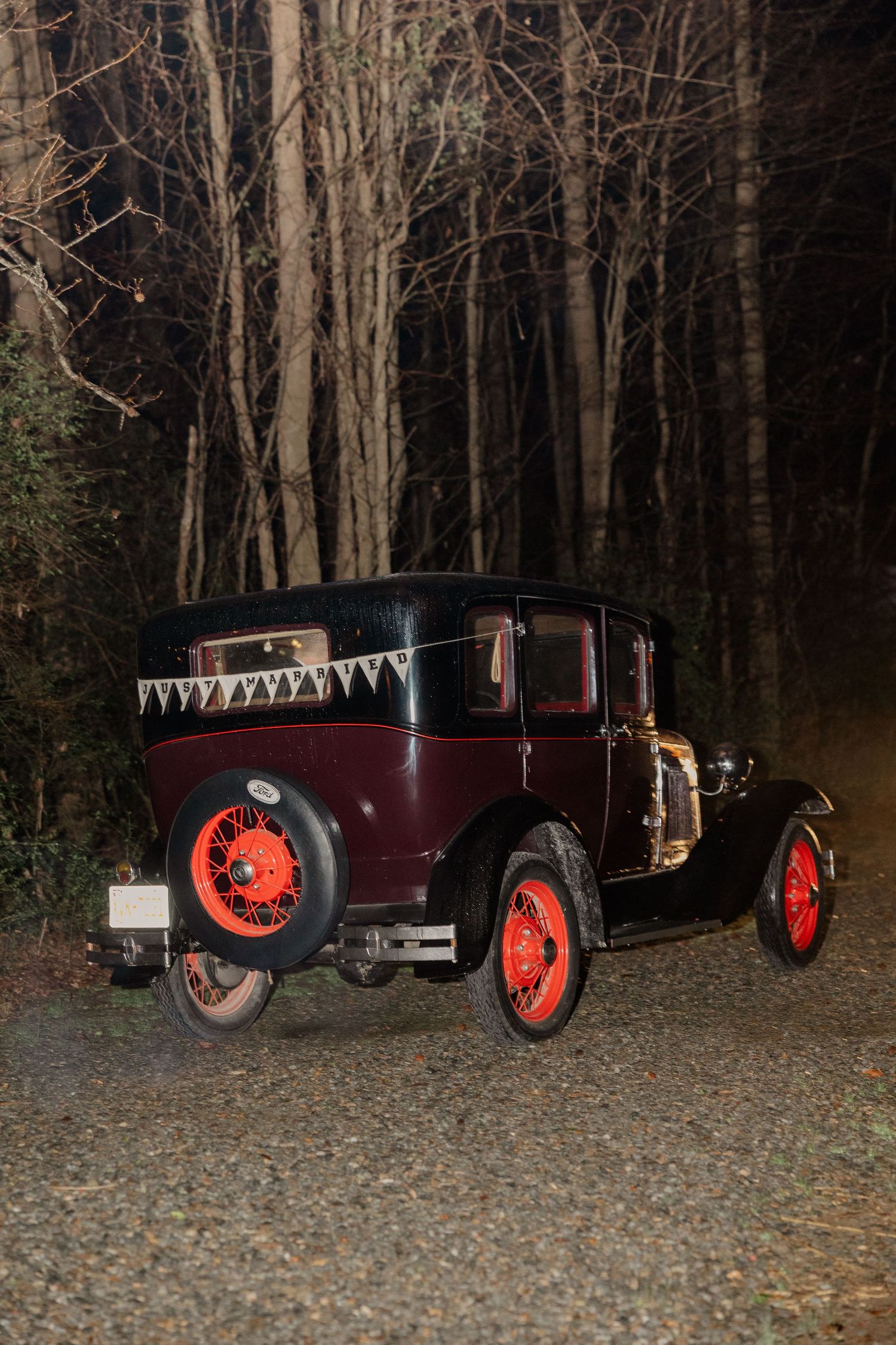 A vintage maroon car with red wheels parked on gravel in front of a dark wooded area.
