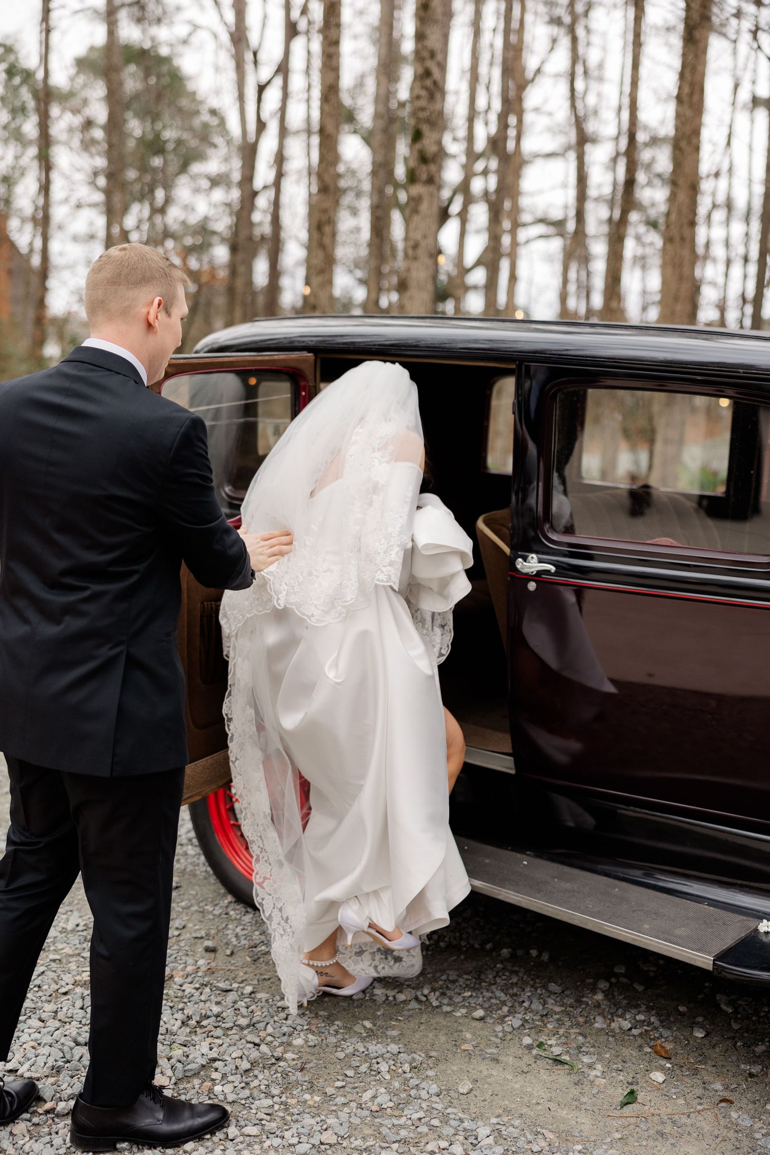 Bride in flowing white dress gets helped into vintage car outside on gravel path.