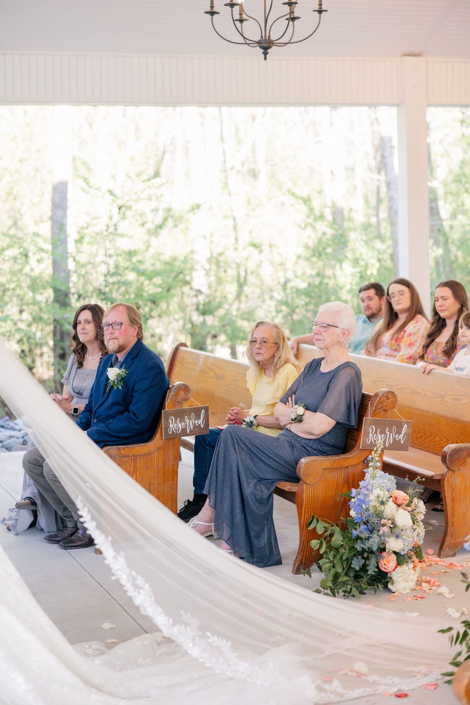 Wedding guests seated on wooden pews in an outdoor venue with flowing white drapery and floral arrangements.