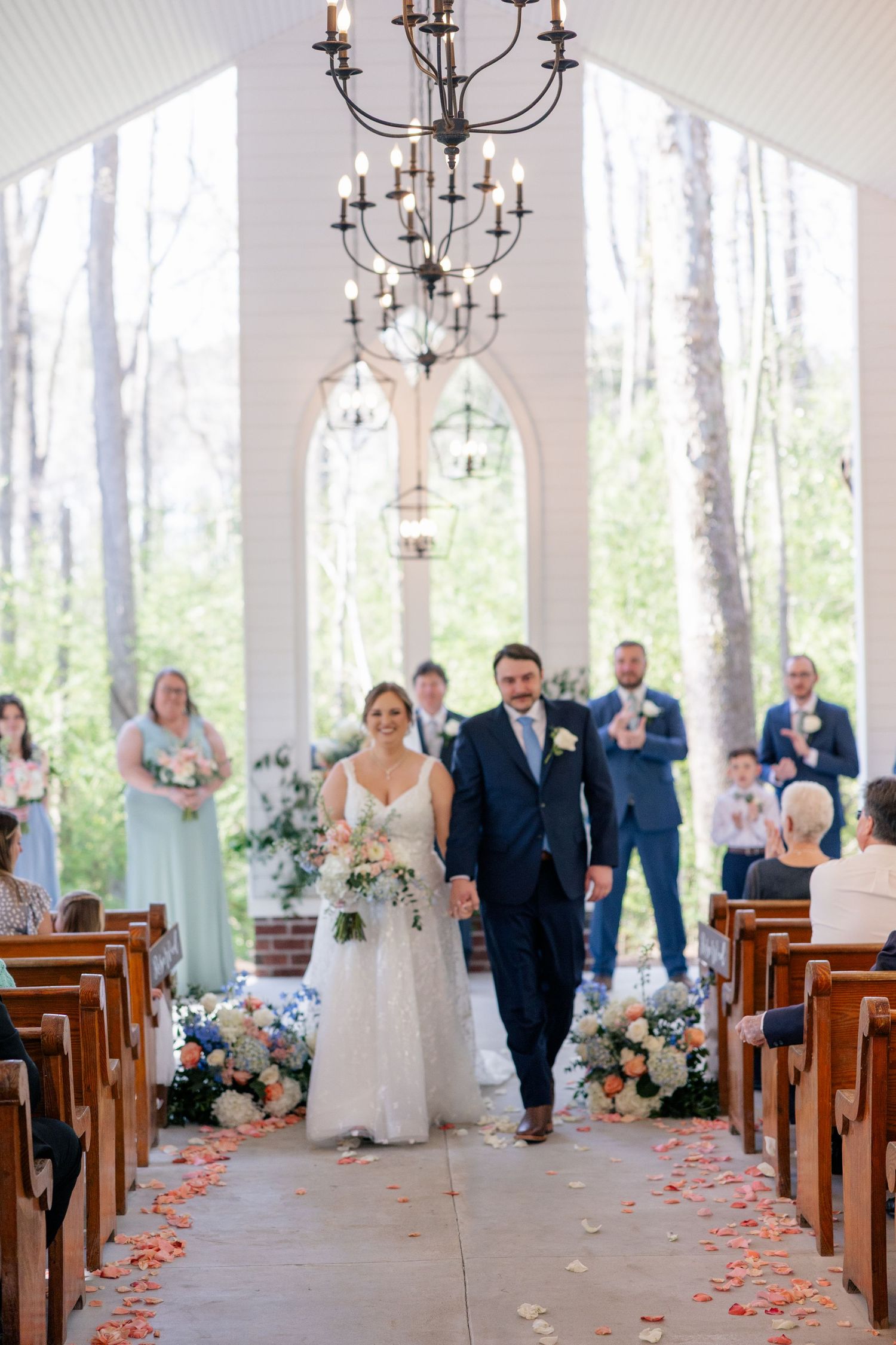 Wedding ceremony procession in a bright white chapel with wooden pews and floral decorations.