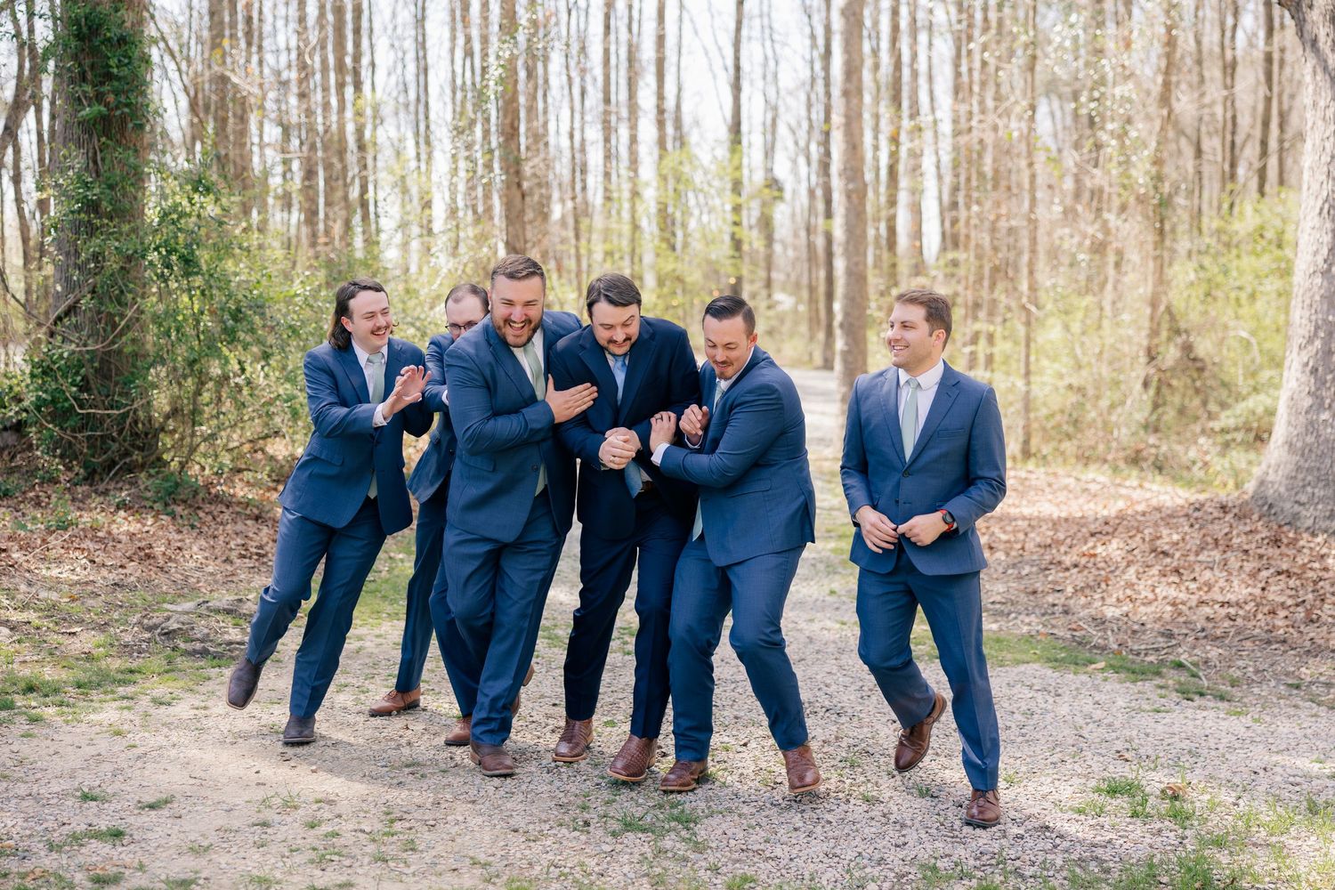 A group in navy suits poses playfully outdoors against a wooded backdrop.