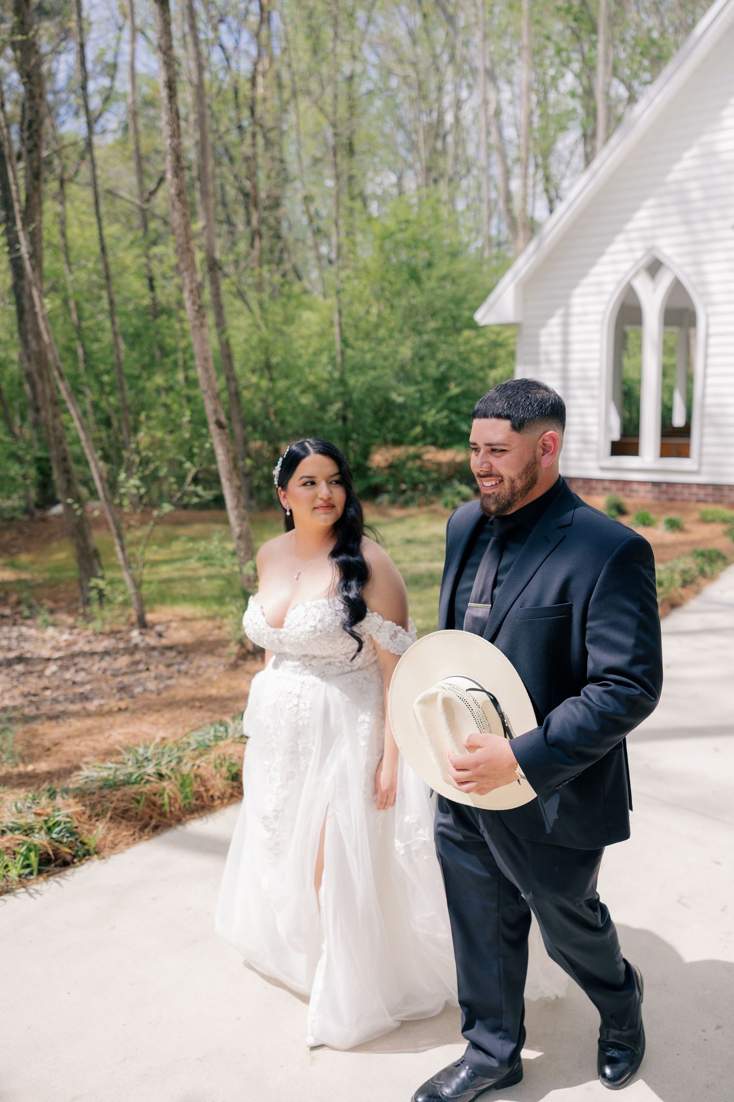 A couple shares a romantic moment outside a white chapel surrounded by trees.