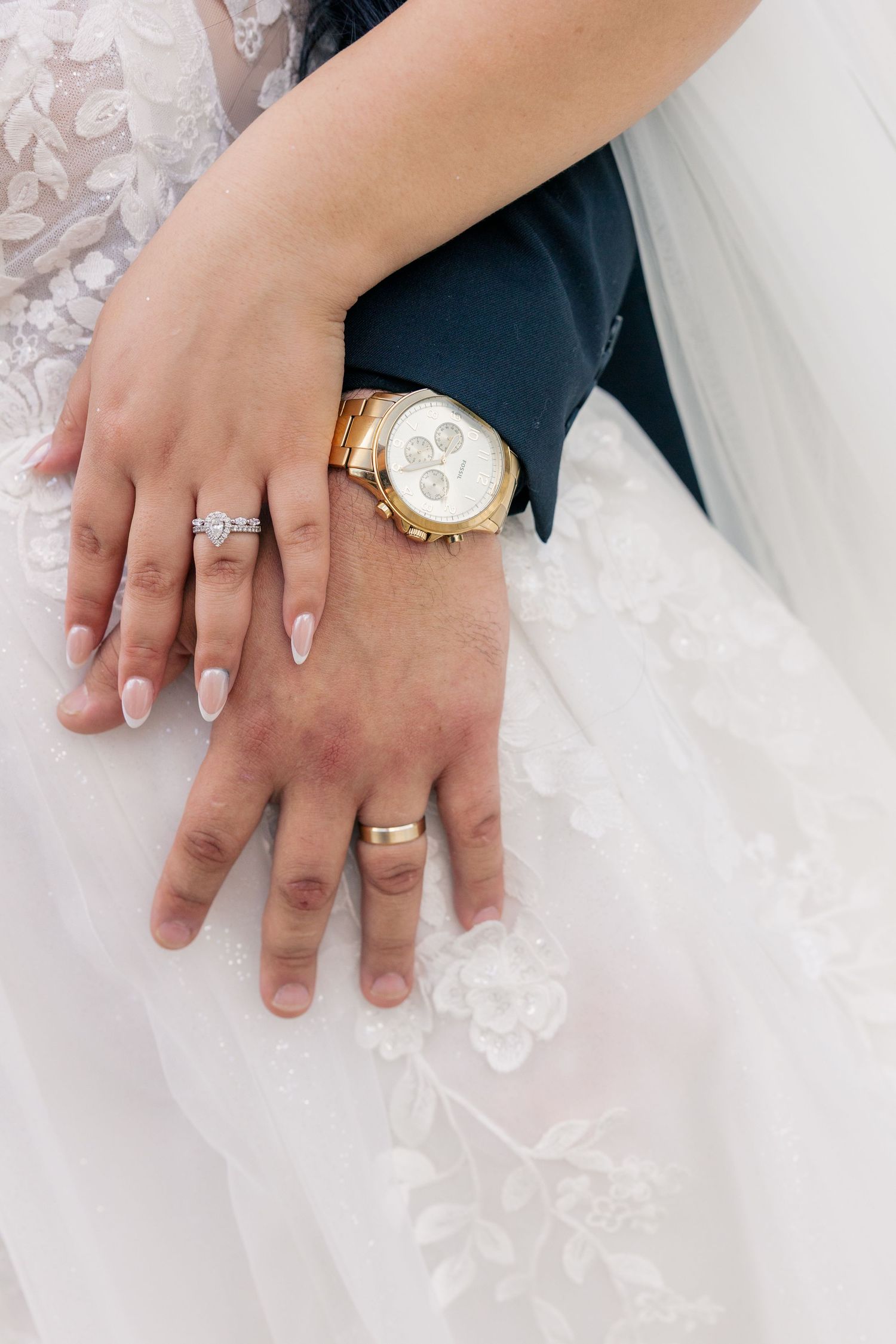 Close-up of wedding rings and jewelry on hands against white lace dress fabric.