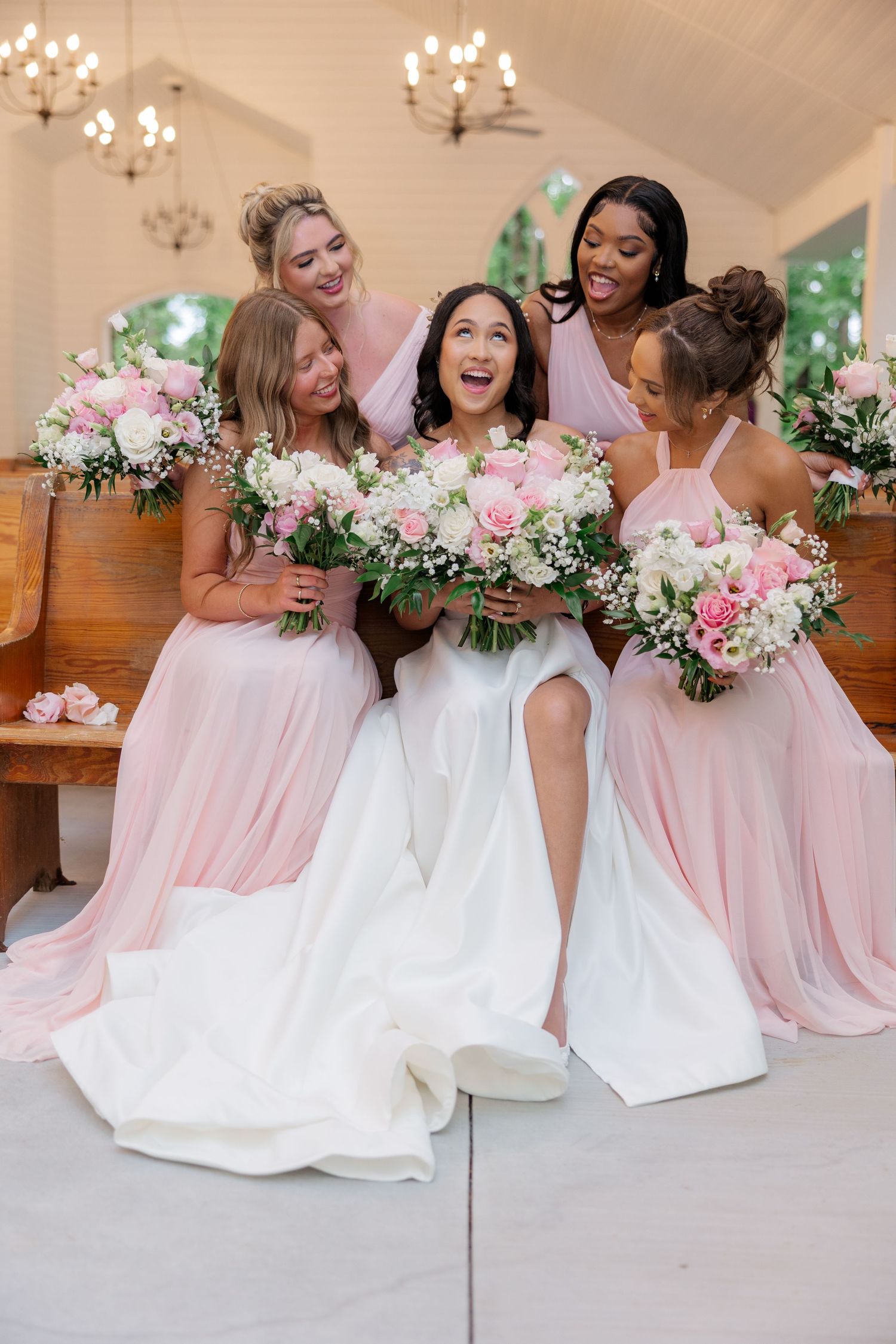 Bridesmaids in pink dresses sit together holding pink and white floral bouquets.