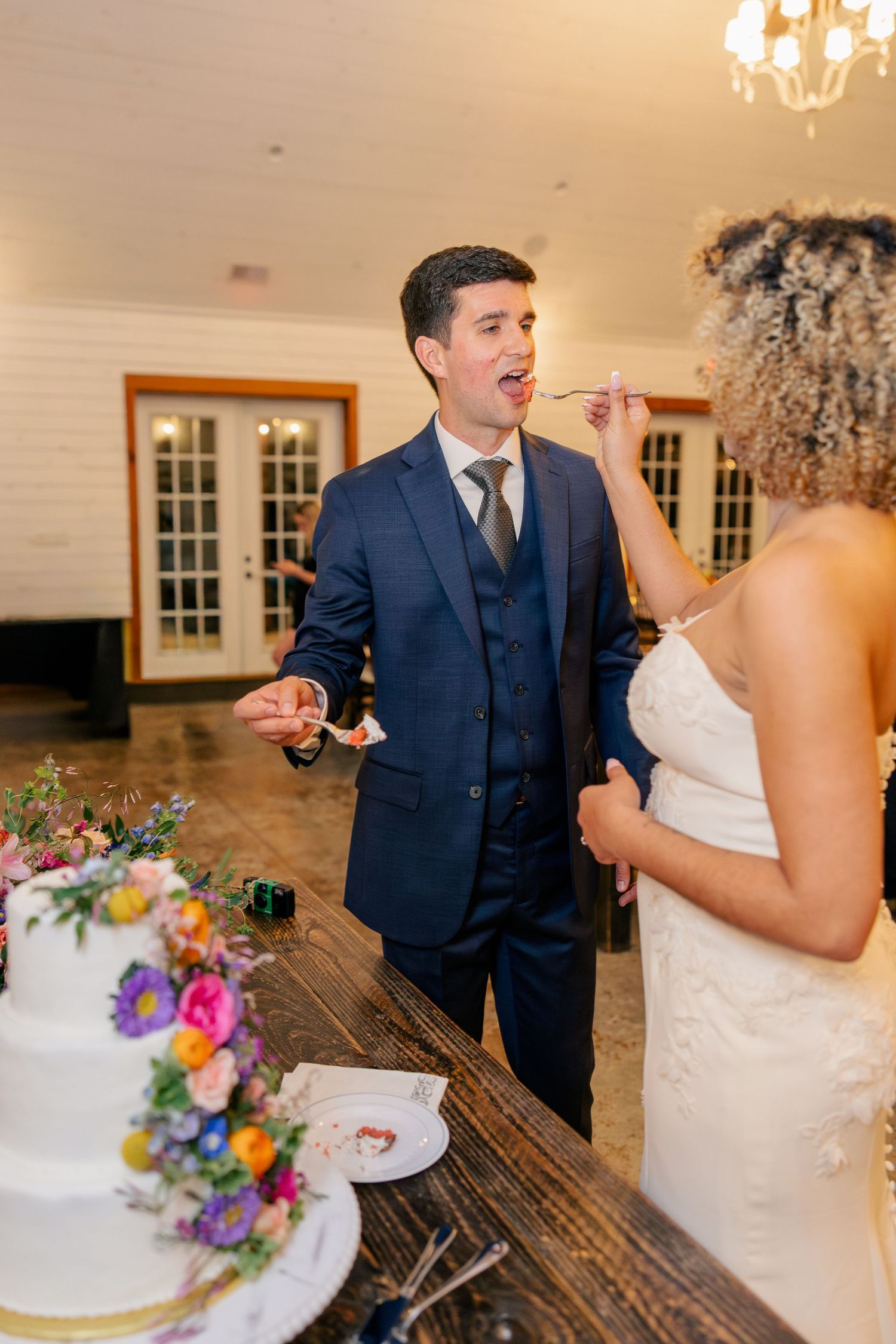 Bride feeding groom wedding cake slice at colorful floral decorated reception table.