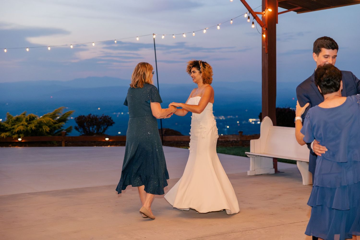 Bride dancing with a guest at an outdoor wedding reception under string lights at dusk with scenic views.
