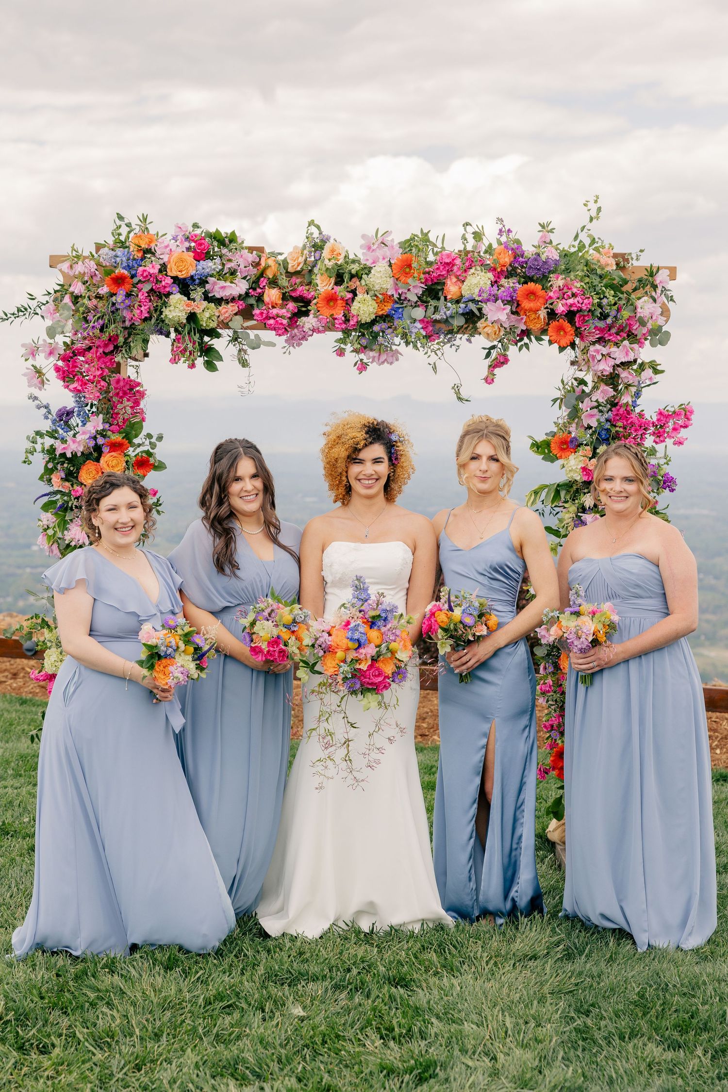 Bride with four bridesmaids in blue dresses holding colorful bouquets under a vibrant floral arch outdoors.
