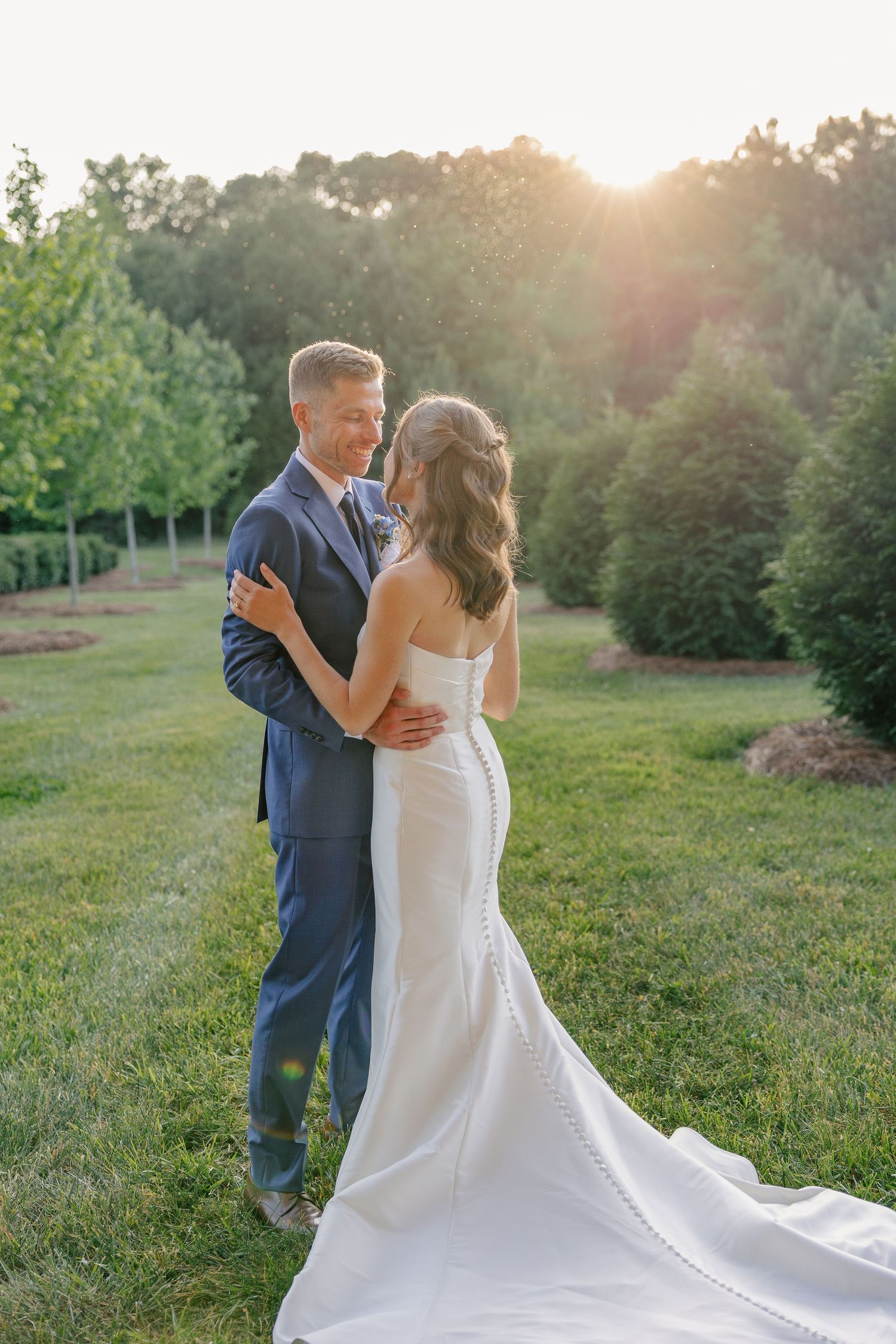 Bride and groom embrace at golden hour sunset surrounded by lush green trees and manicured garden landscape.