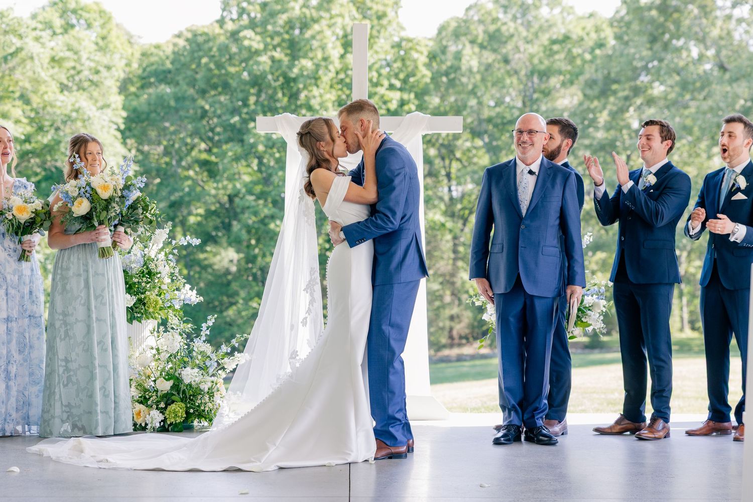 Bride and groom share first kiss at outdoor wedding ceremony with bridal party in blue and sage green.