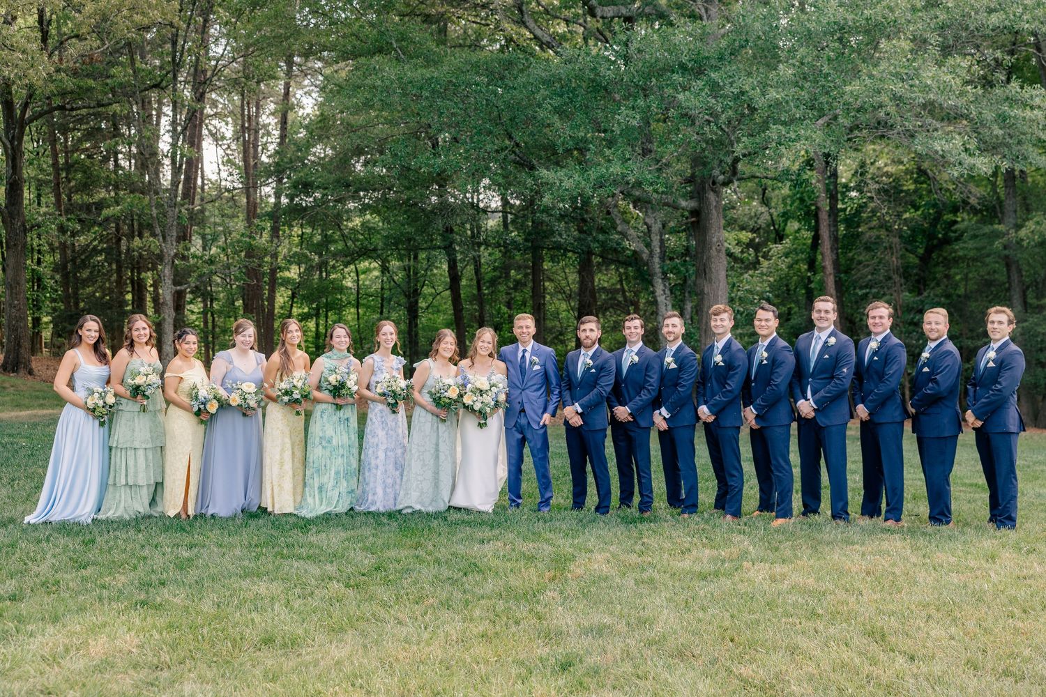 Wedding party in blue suits and pastel bridesmaid dresses poses in a wooded outdoor setting.