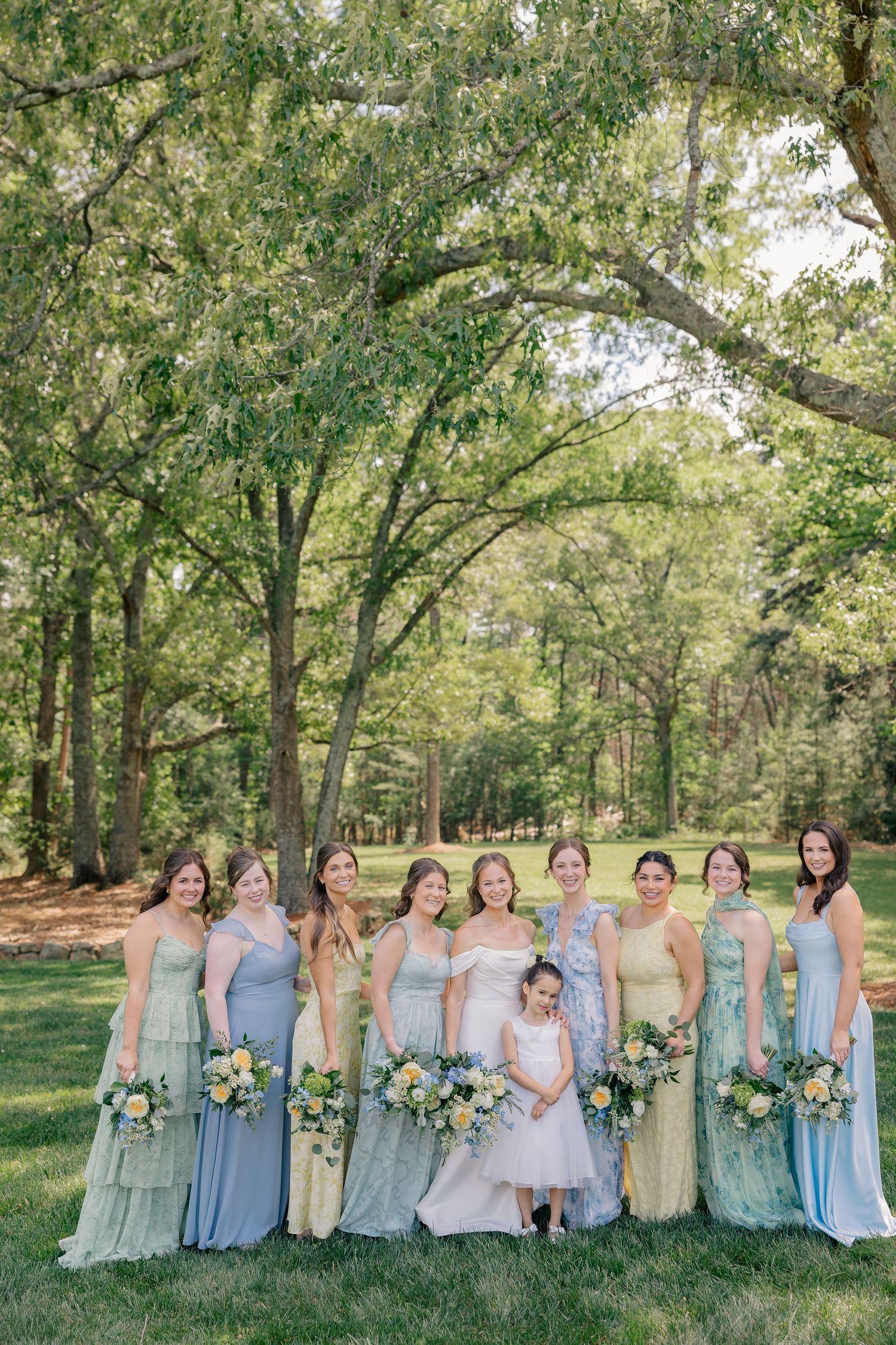 Bridesmaids in mismatched blue and yellow gowns pose with bride under lush green trees.