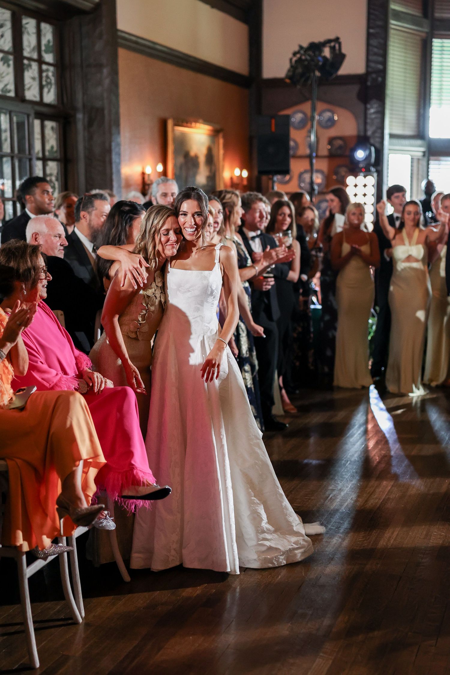Bride in white gown smiles and hugs a guest during wedding reception in elegant wood-paneled venue.