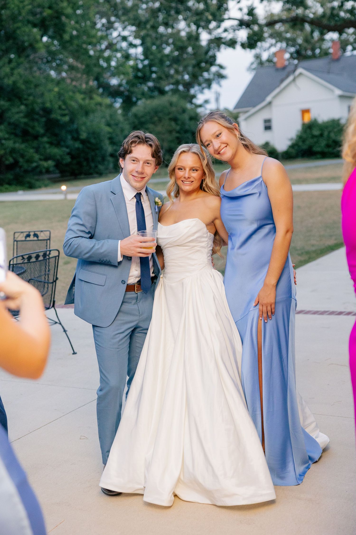 Bride in strapless gown poses with groom in blue suit and bridesmaid at outdoor reception.