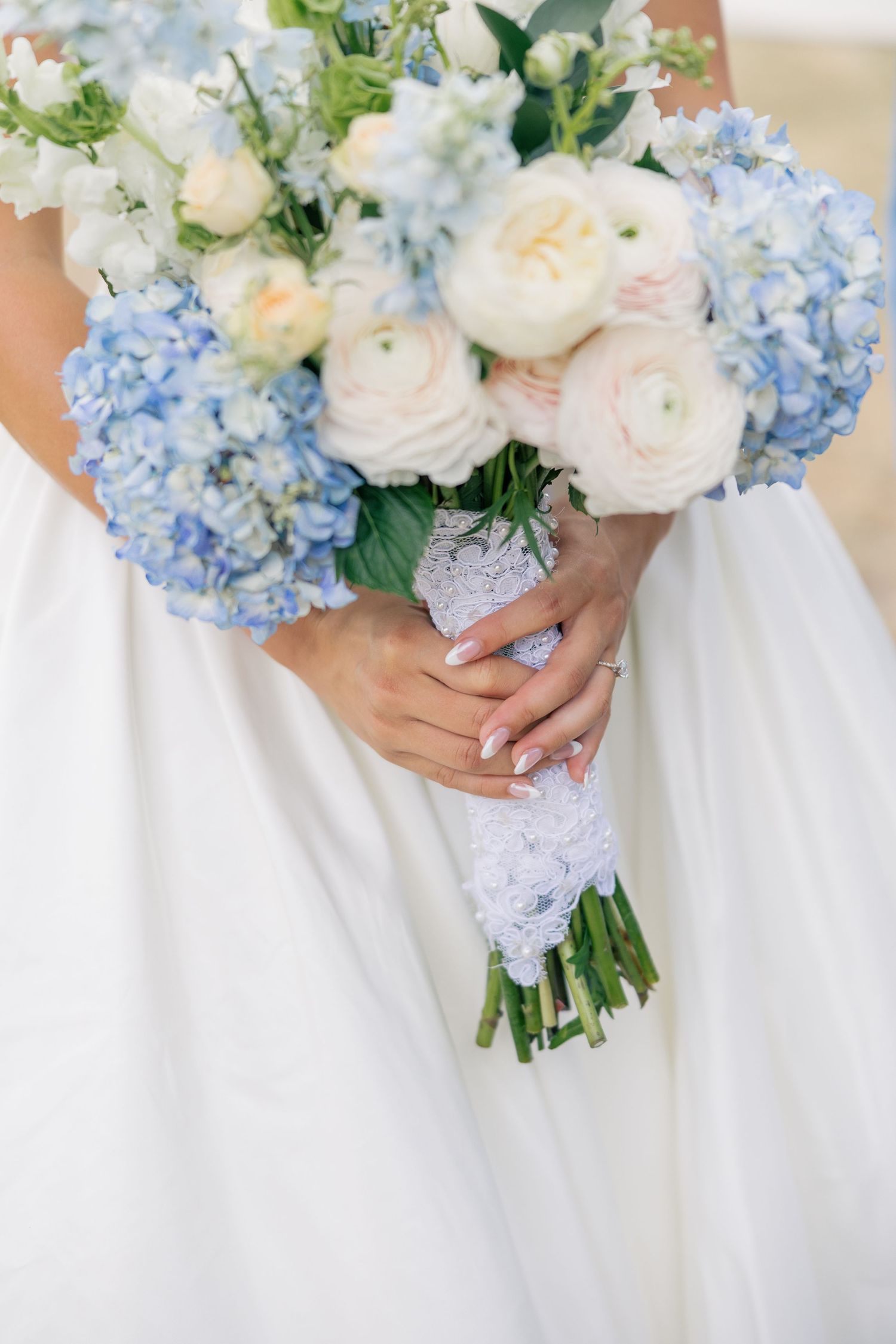 Bride holding blue hydrangea and white ranunculus bouquet with lace wrap.