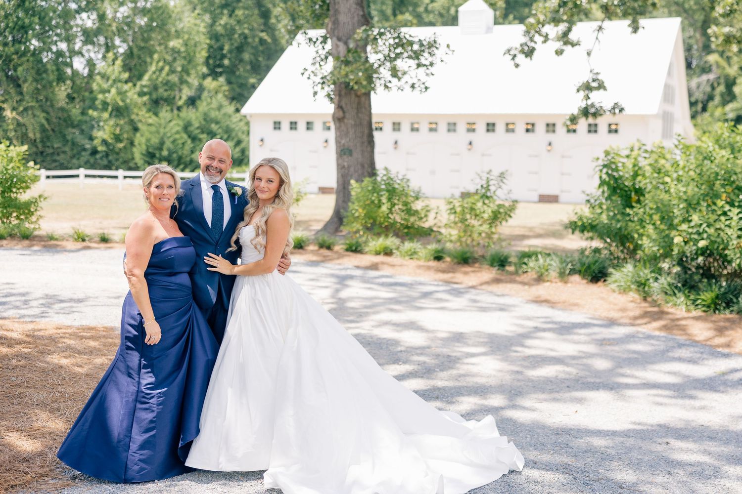 Bride in white gown poses with parents in navy attire outside white barn wedding venue.