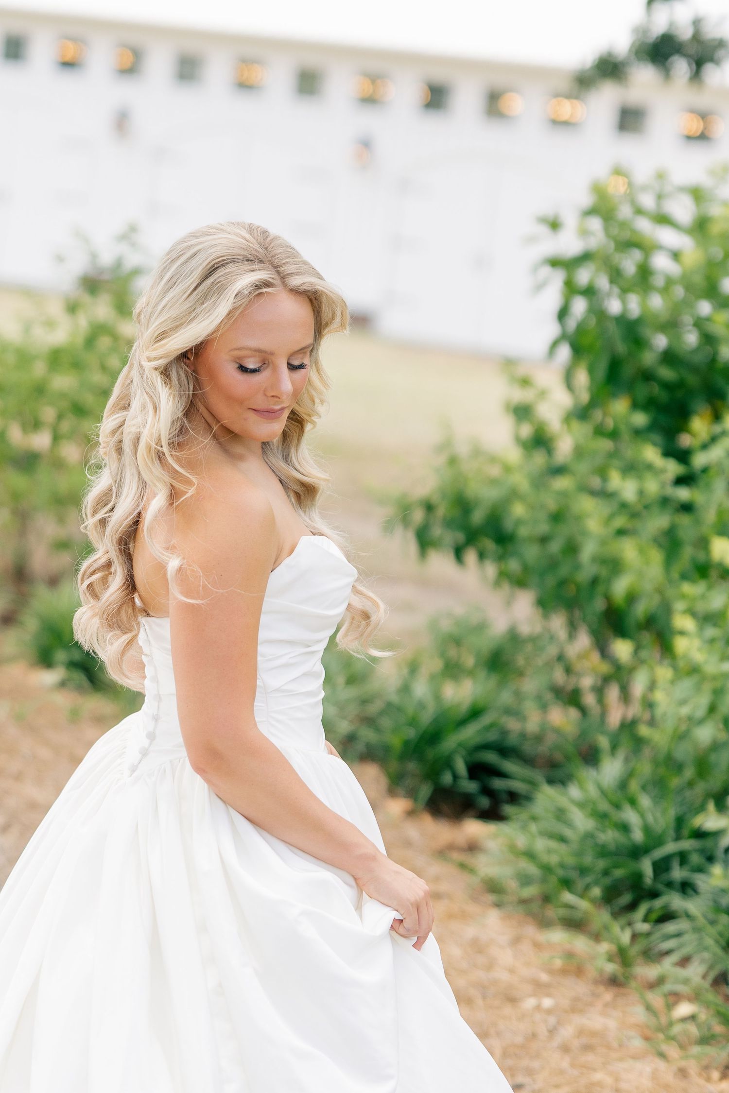Blonde bride in strapless white ballgown twirls in garden near white barn wedding venue.