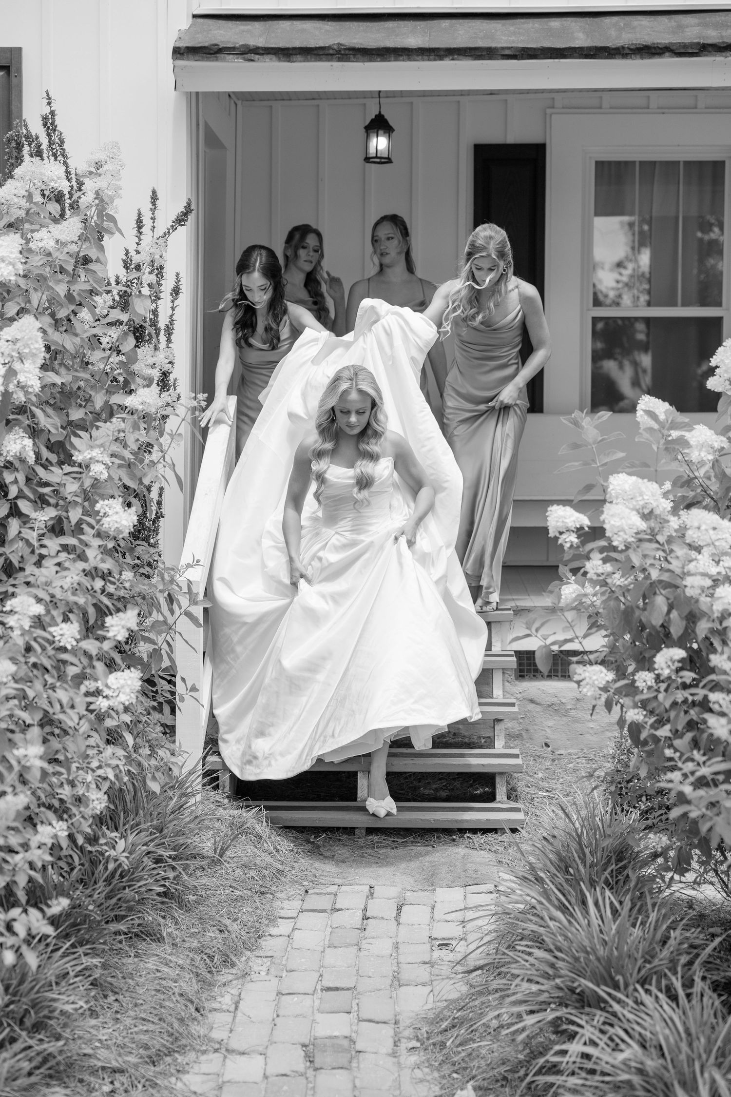 Bride in white gown descends steps with bridesmaids holding her train in black and white wedding photo.