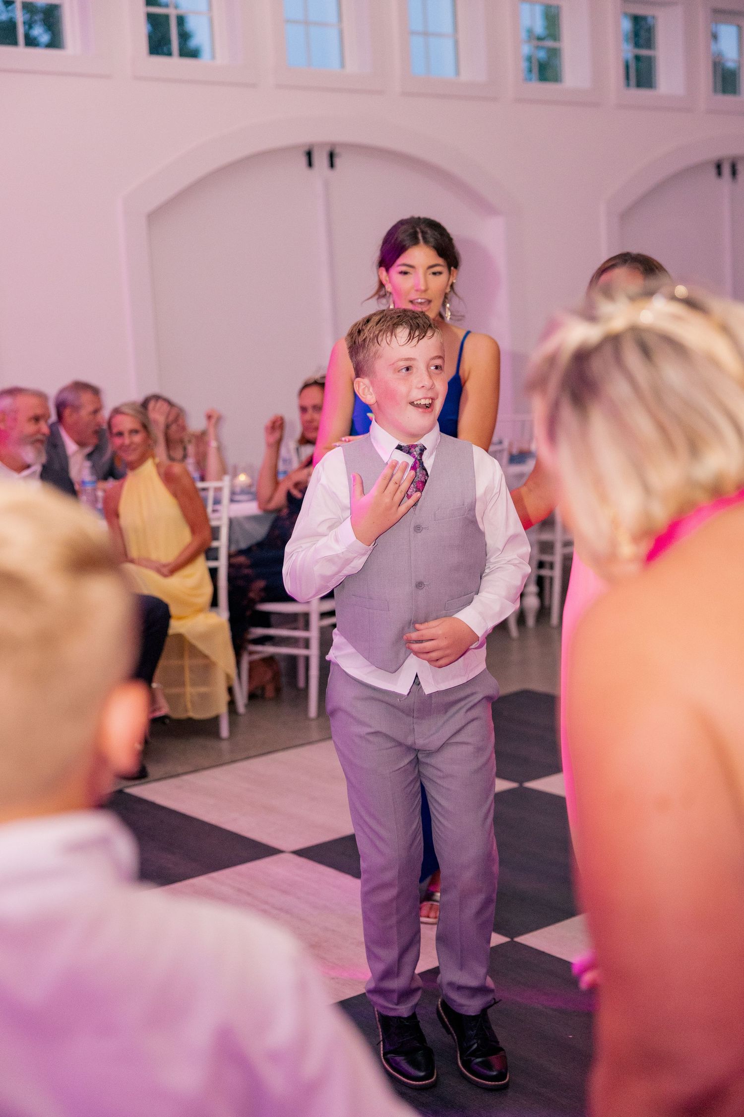 Young boy in gray suit vest dancing at a wedding reception on a checkered dance floor.